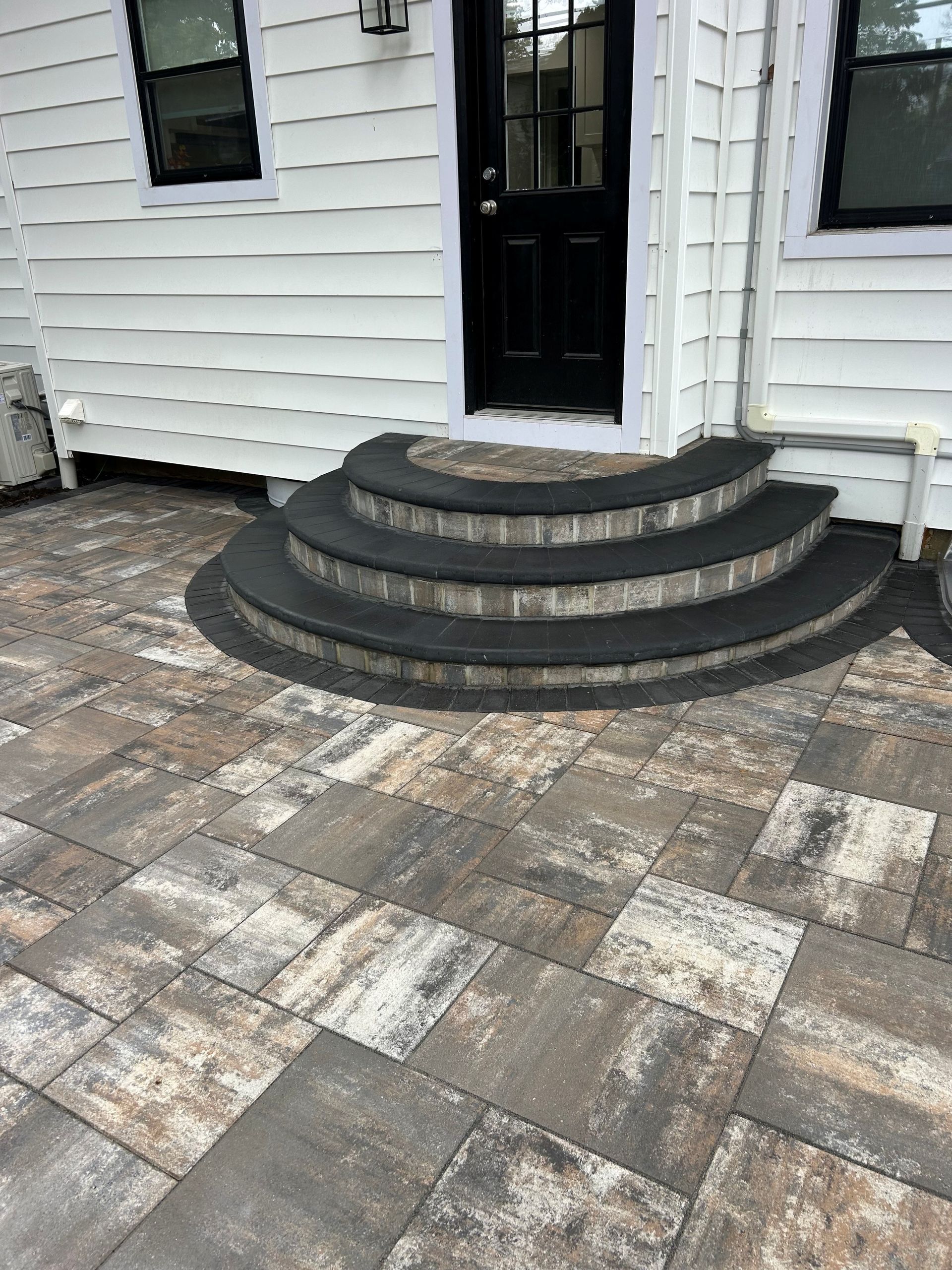 Brick patio with steps leading to a dark door. White siding, neutral pavers, black steps.