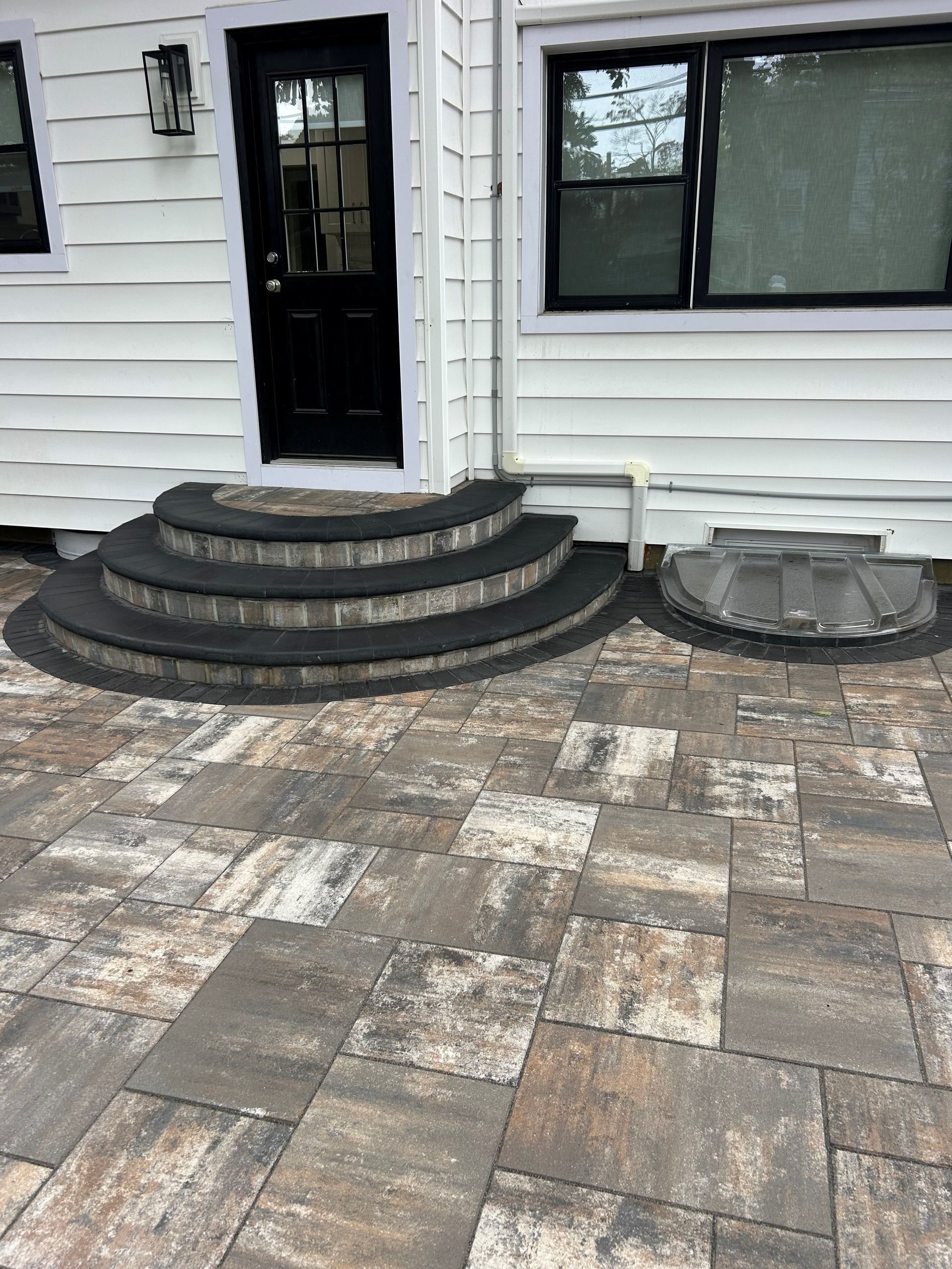 Exterior view: black door, curved steps, patterned pavers, and a window.