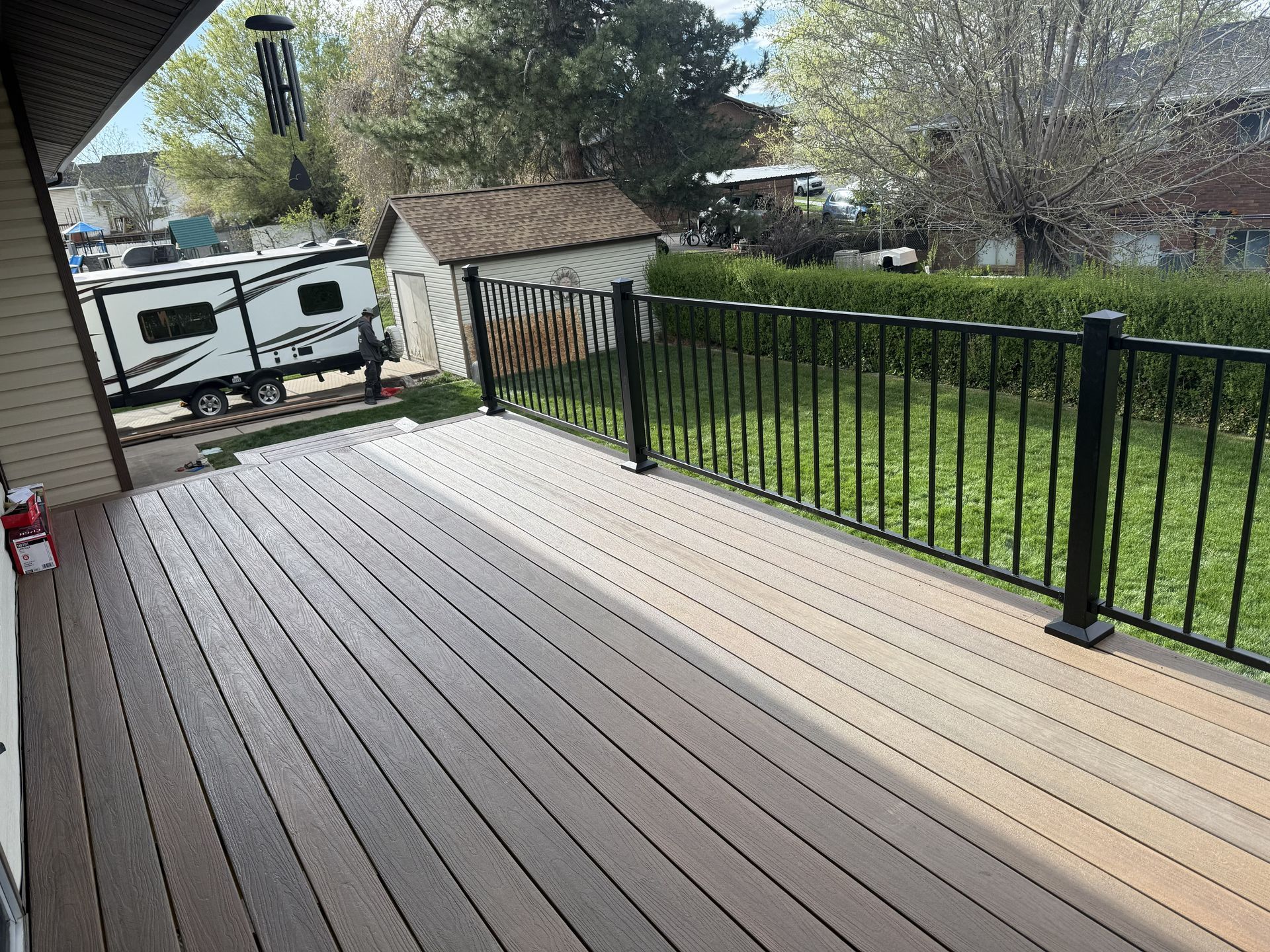 A two-toned composite deck with dark and light brown boards, black metal railings, a shed, and a camper in the background.