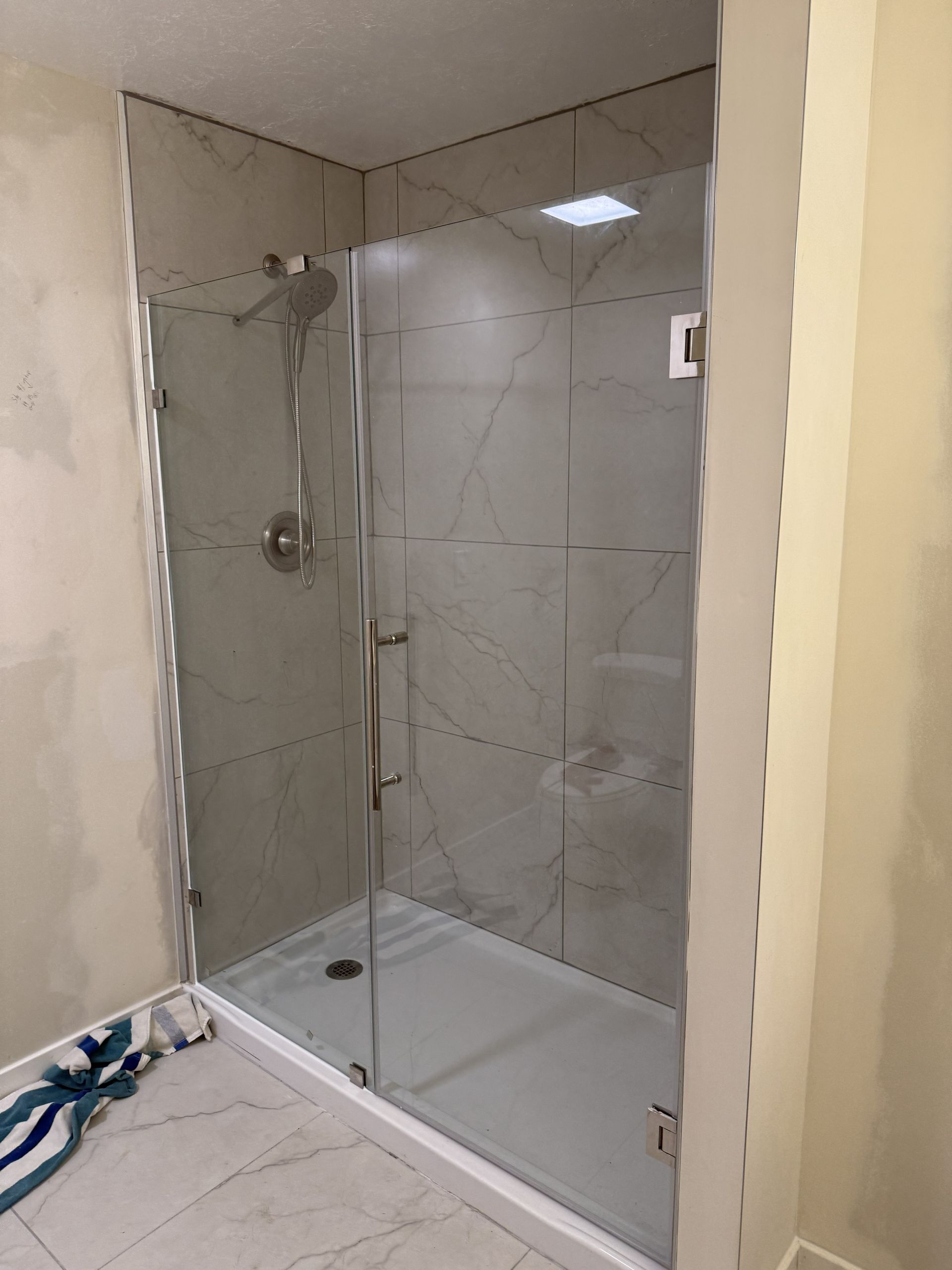A glass shower enclosure with a chrome shower head and light-gray marble-patterned tiles in a bathroom under construction.