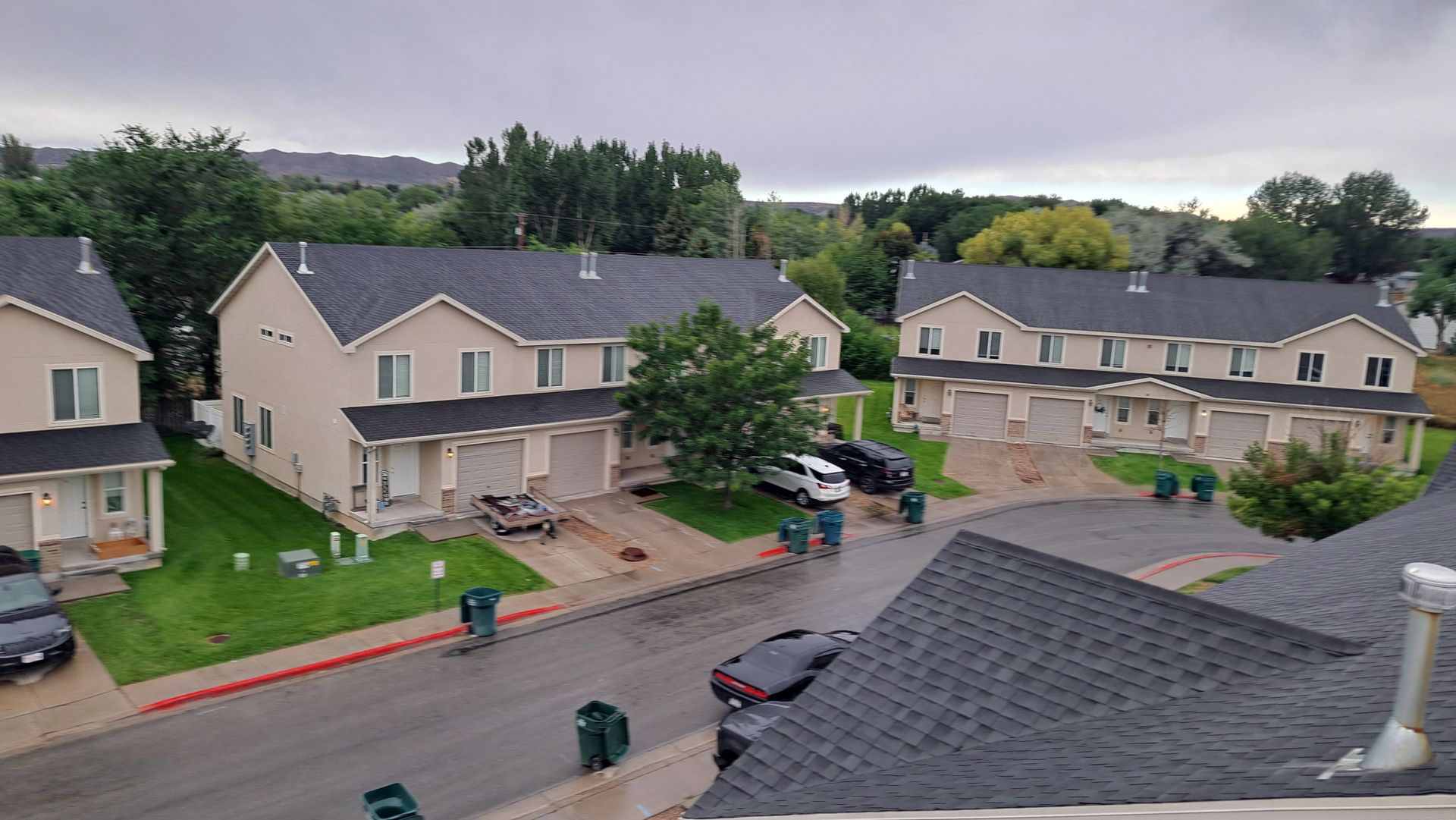 An elevated view of beige townhouses with dark shingled roofs, green lawns, and a quiet street on an overcast day.