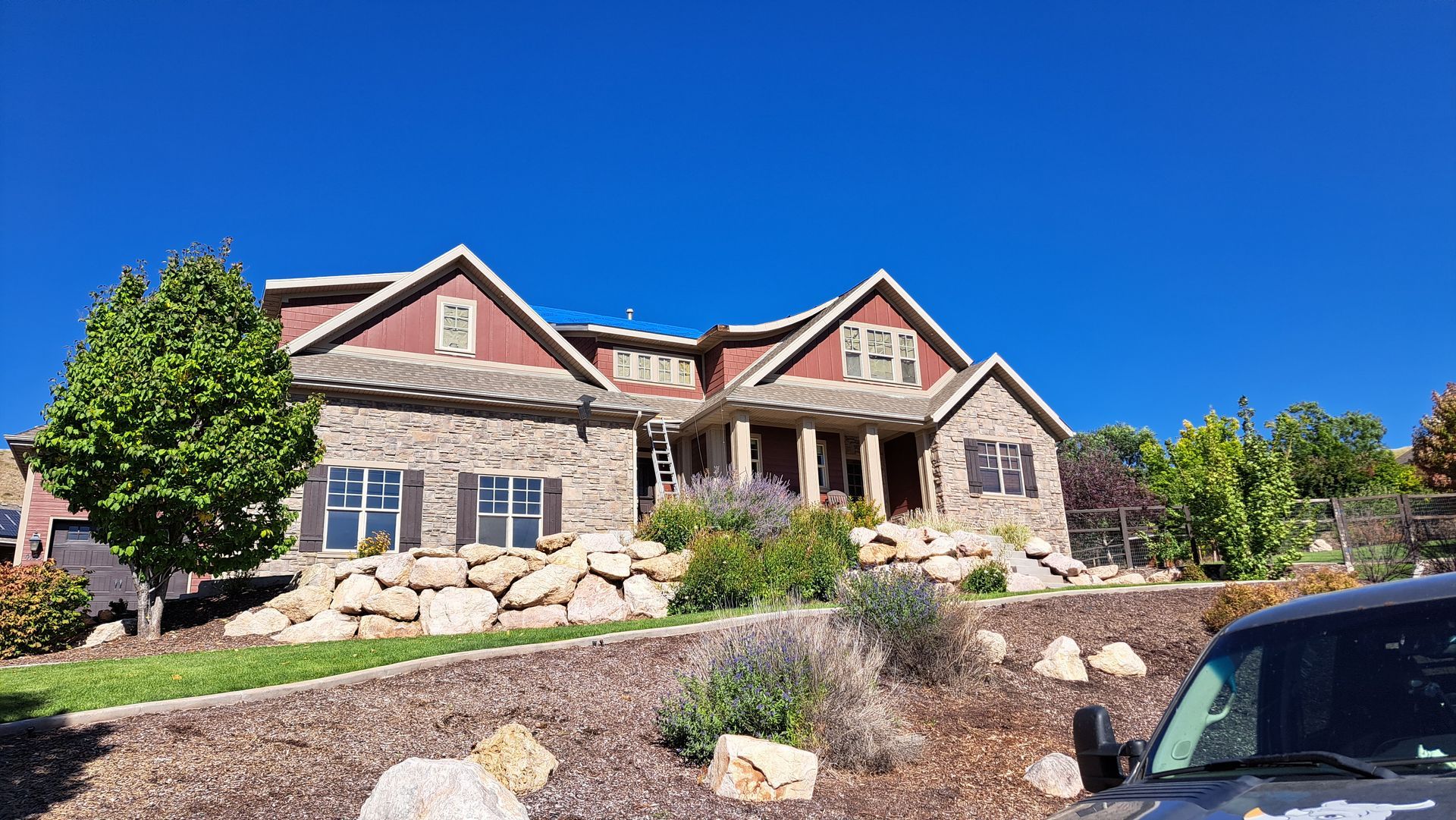 A two-story stone and reddish-brown house sits on a hill above a landscaped yard with rocks and a green tree.