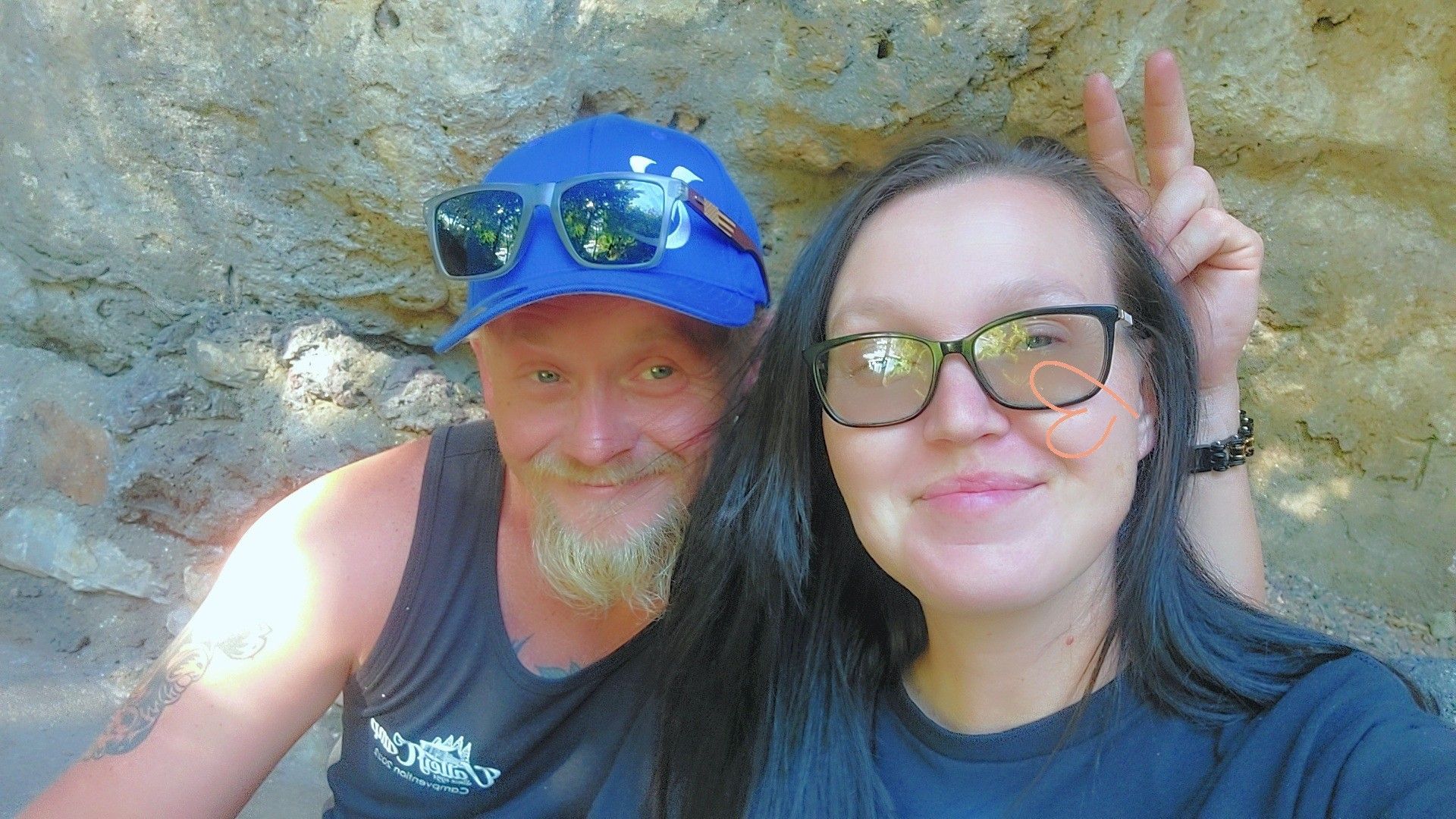 A person in a blue cap and sunglasses and a person with dark hair and glasses pose together in front of a rock wall.