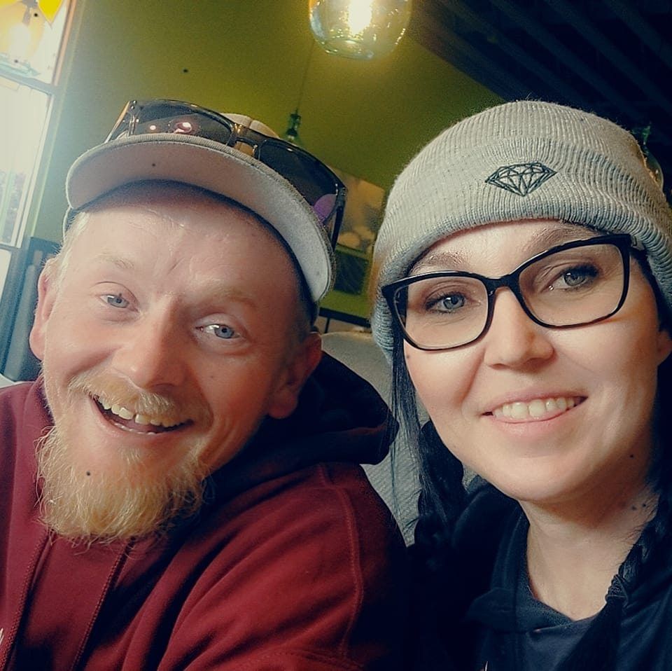 A person in a baseball cap and a person in a beanie with a diamond logo smiling together in an indoor setting.