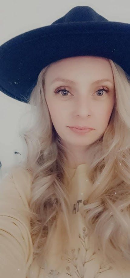 A person with blonde hair and a neutral expression wearing a dark fedora and a light-colored long-sleeved shirt.
