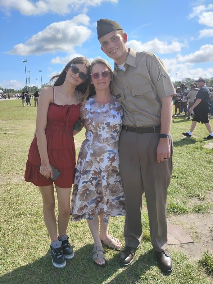 Three people stand together outdoors on a sunny, grassy field. A person in a military uniform is on the right.