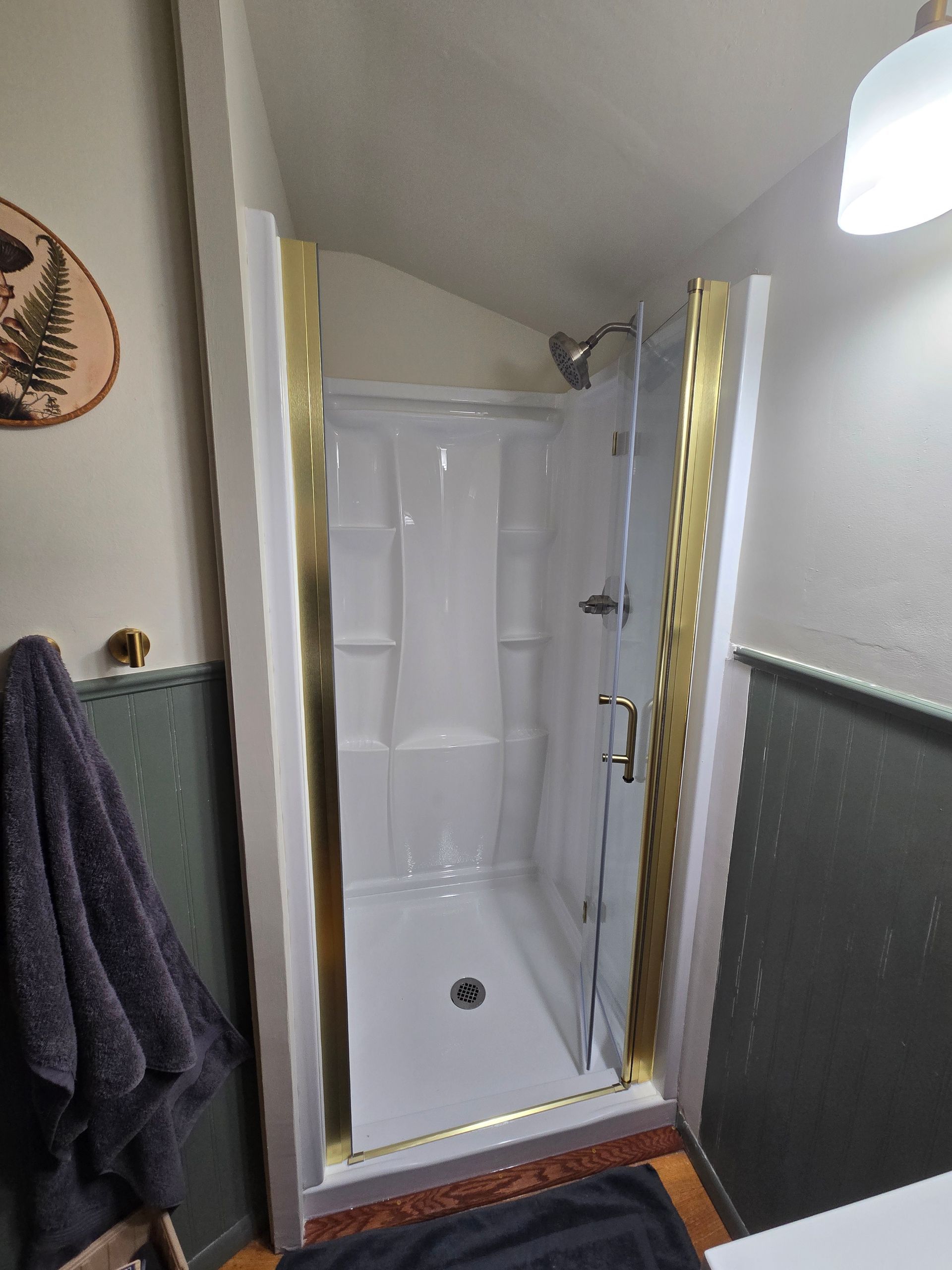 A shower stall with white walls and a gold-trimmed door, set in a bathroom with dark green wall paneling.