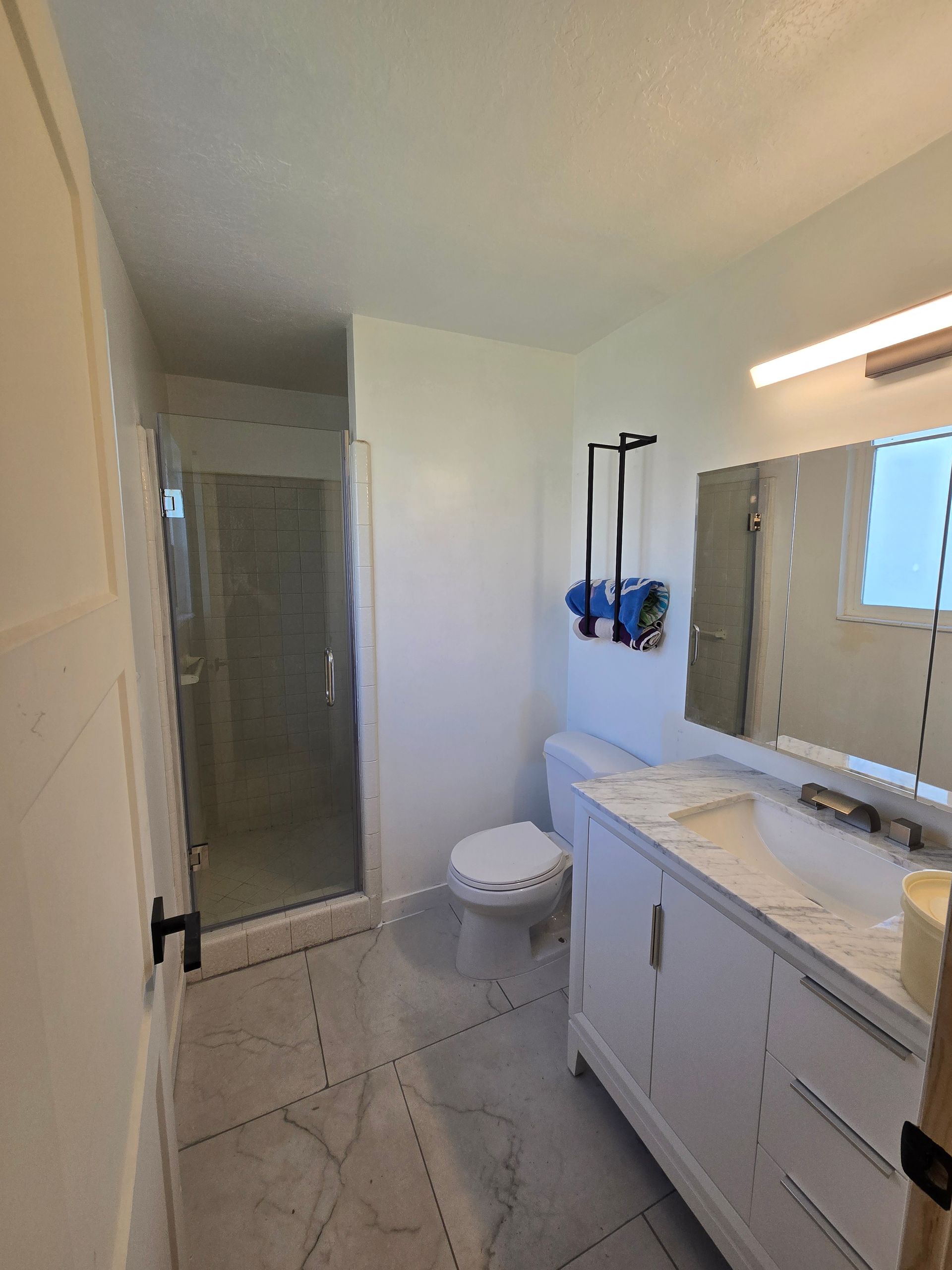 A bright, modern bathroom with a glass-enclosed shower, a white vanity with a marble top, and a toilet against a white wall.