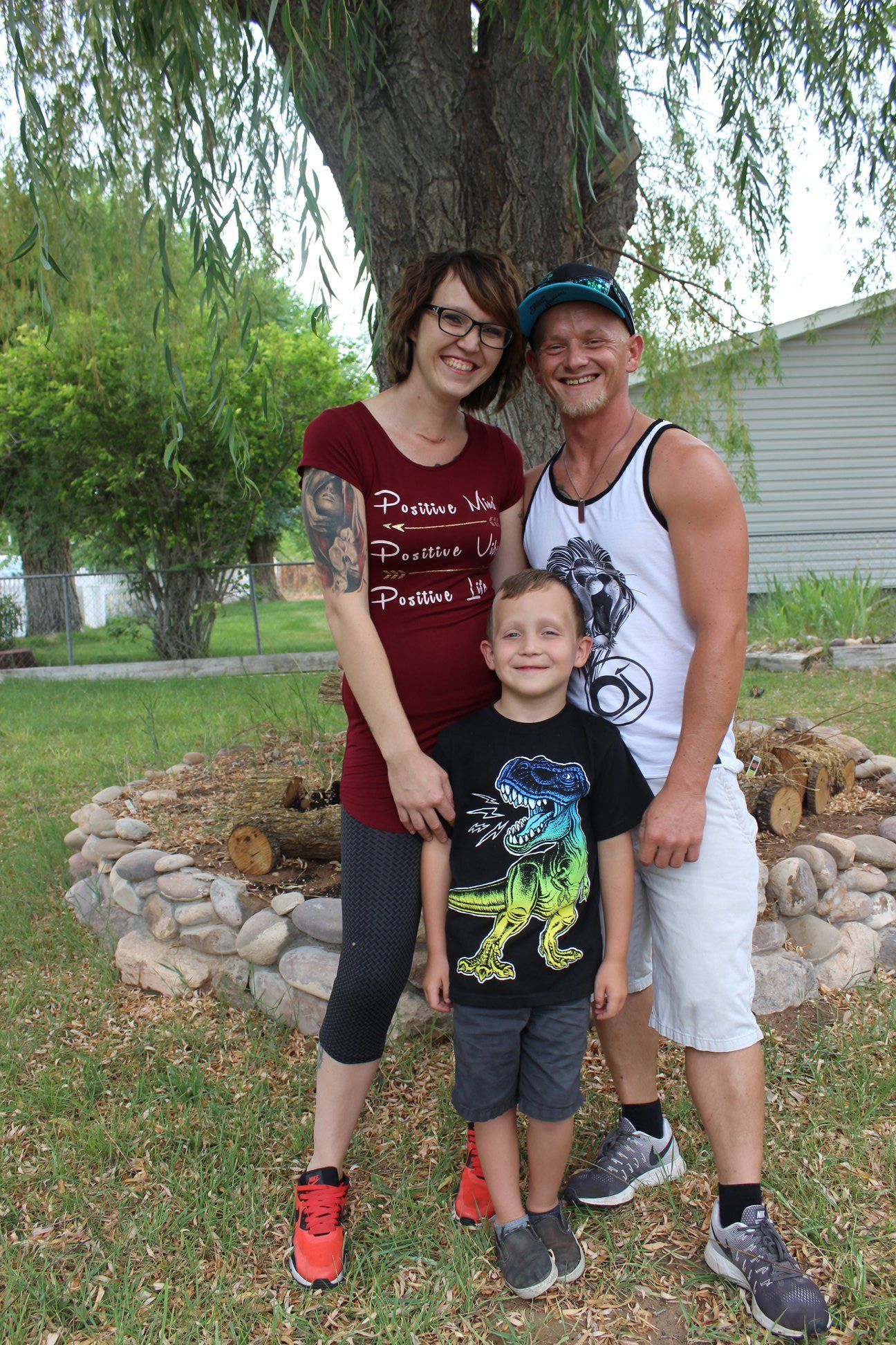 A family of three stands together smiling in front of a tree and a stone-lined garden bed outdoors on a sunny day.