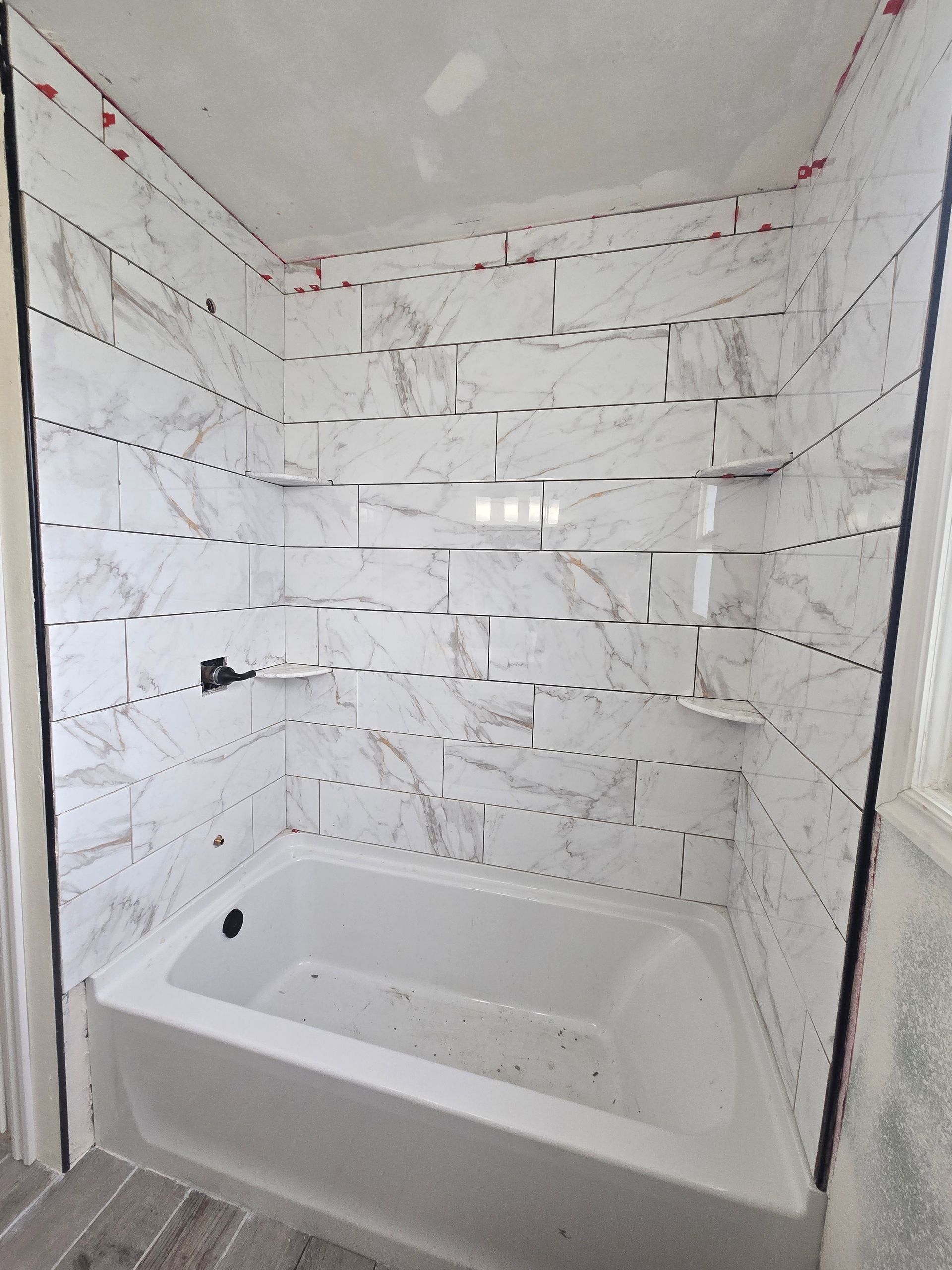 A bathroom shower under construction with white marble-patterned tiles installed above a white bathtub.