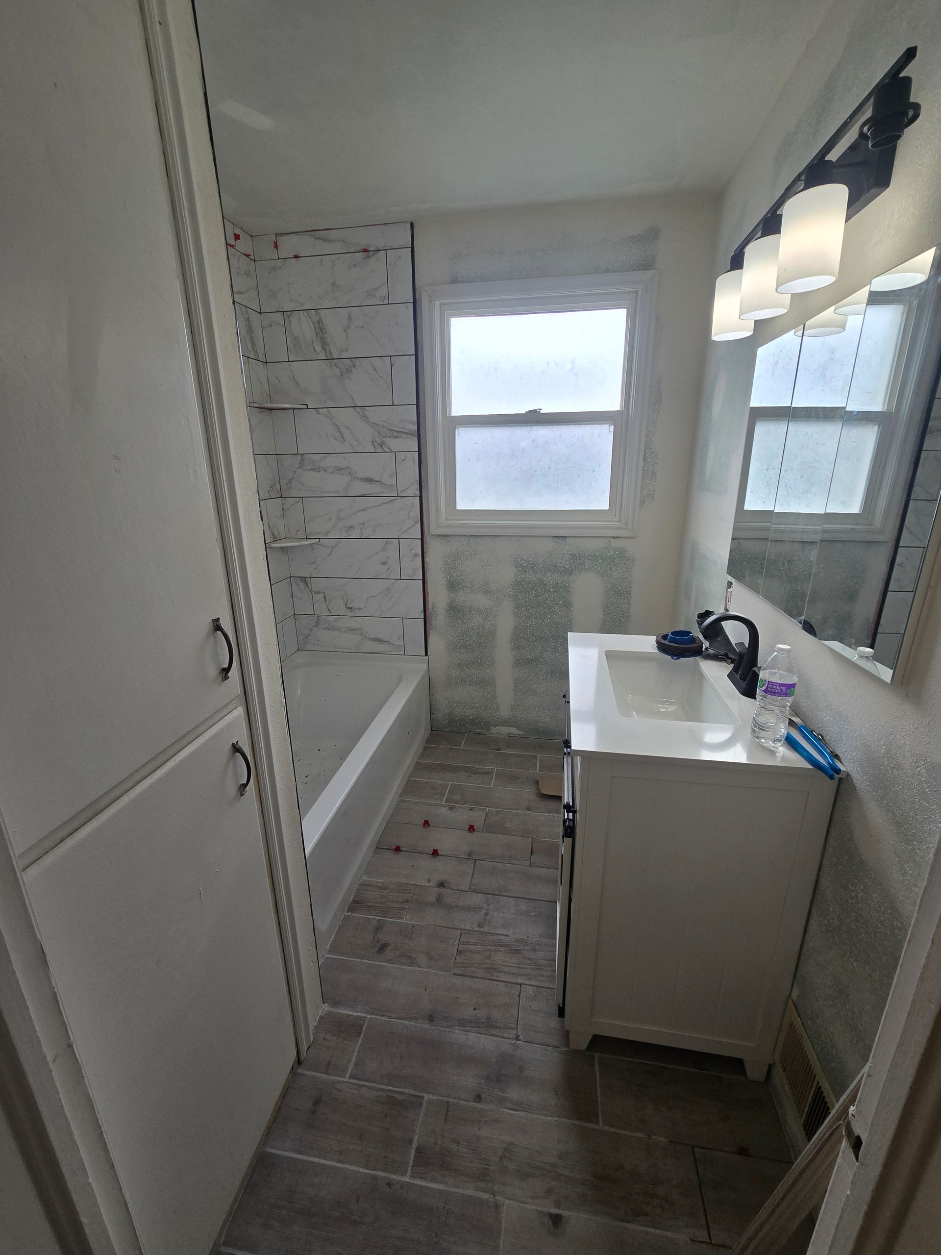 A narrow bathroom featuring white cabinets, a vanity with a dark faucet, a tiled floor, and a tub with stone-like walls.