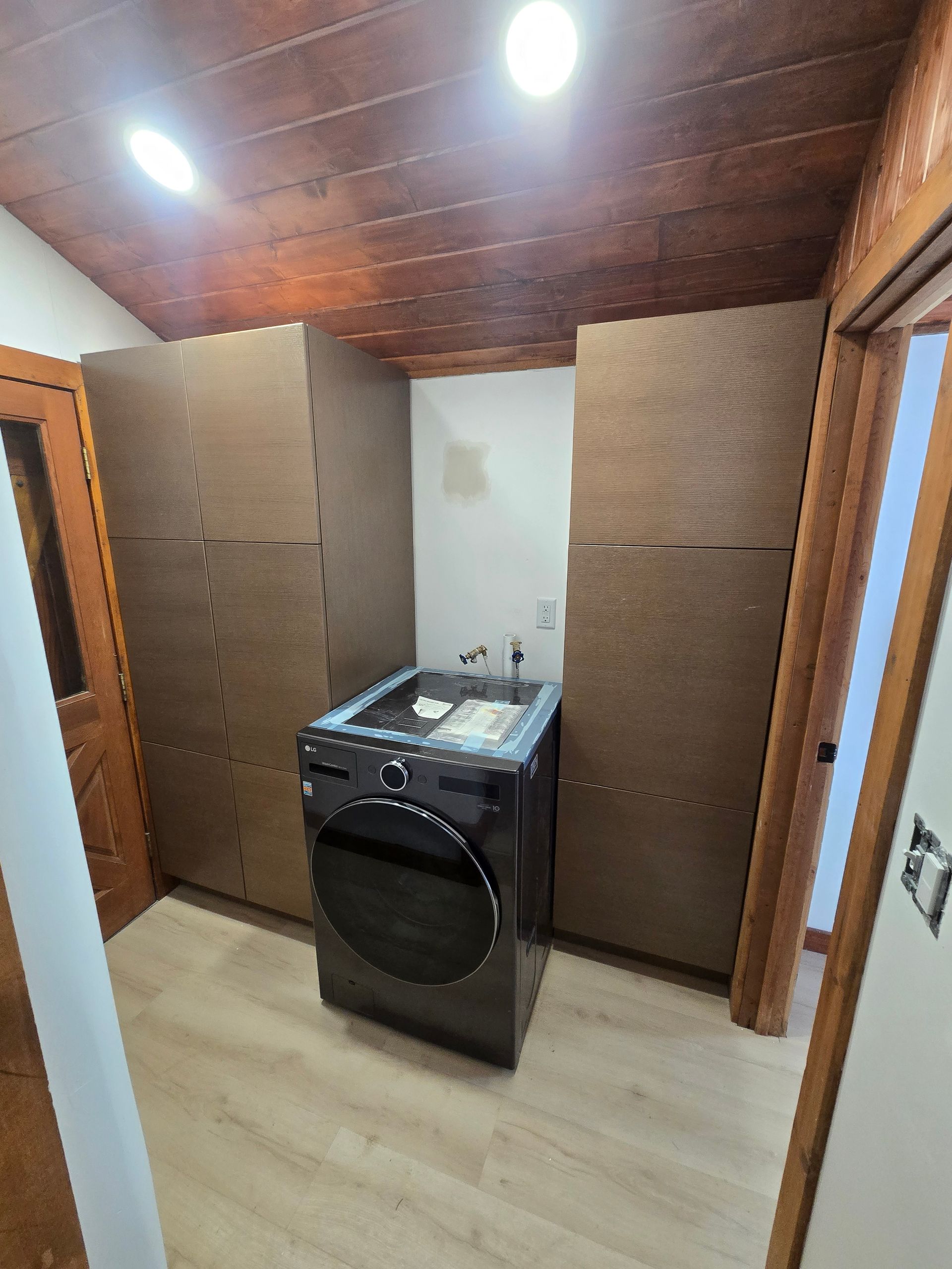 A dark gray washing machine sits between two tall, wood-finished storage cabinets in a room with a wooden ceiling.