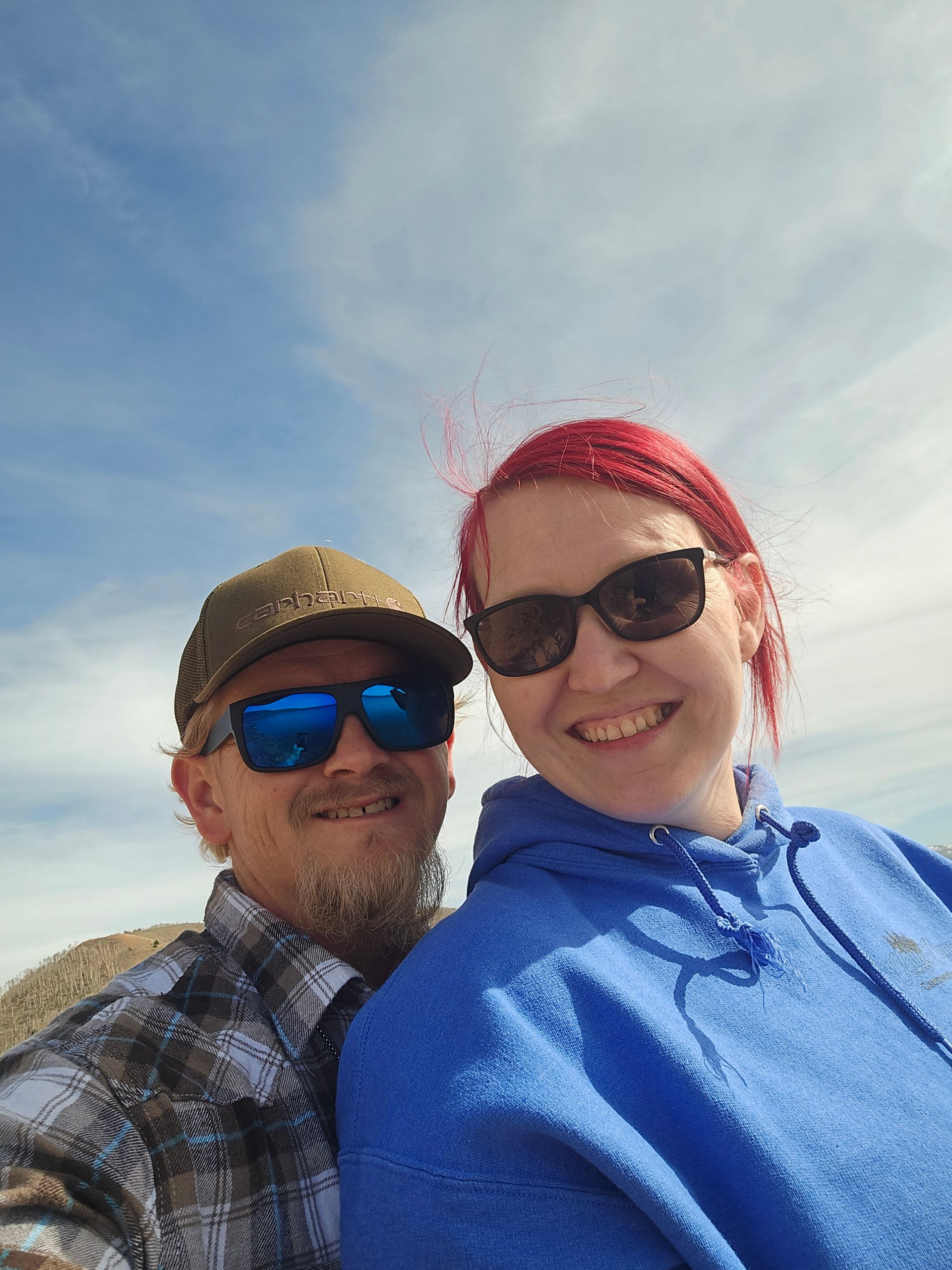 A smiling couple wearing sunglasses poses for a selfie outdoors against a bright blue sky.