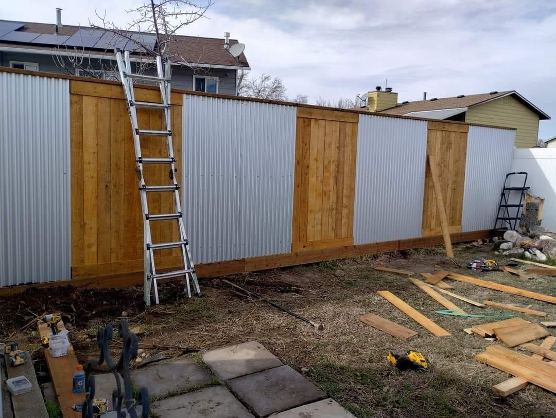 A metal and wood privacy fence under construction in a backyard, with a tall silver ladder leaning against it.