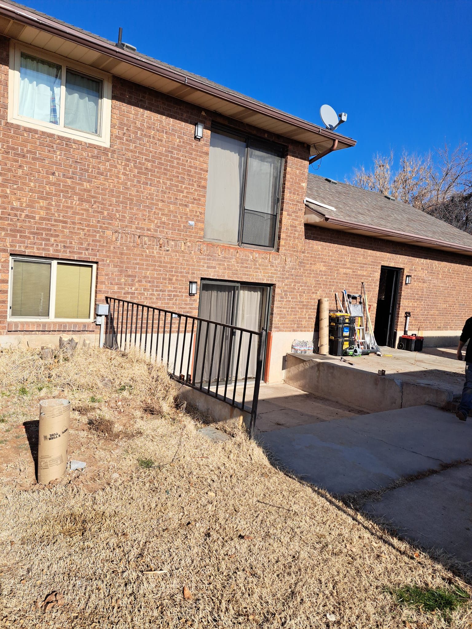 A two-story brick house with a gravel-covered yard and a black metal railing leading to a patio area under a clear sky.