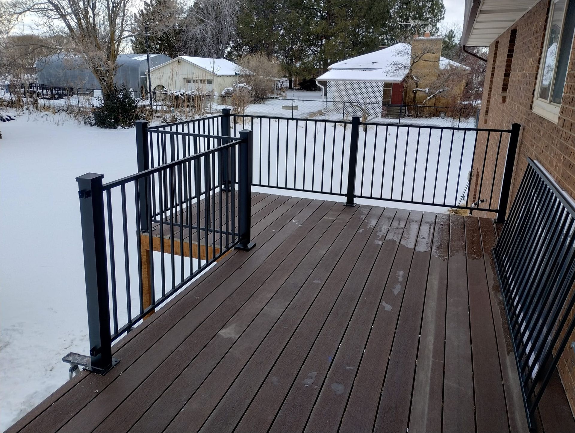 A brown wooden deck with black metal railings overlooking a snow-covered backyard.