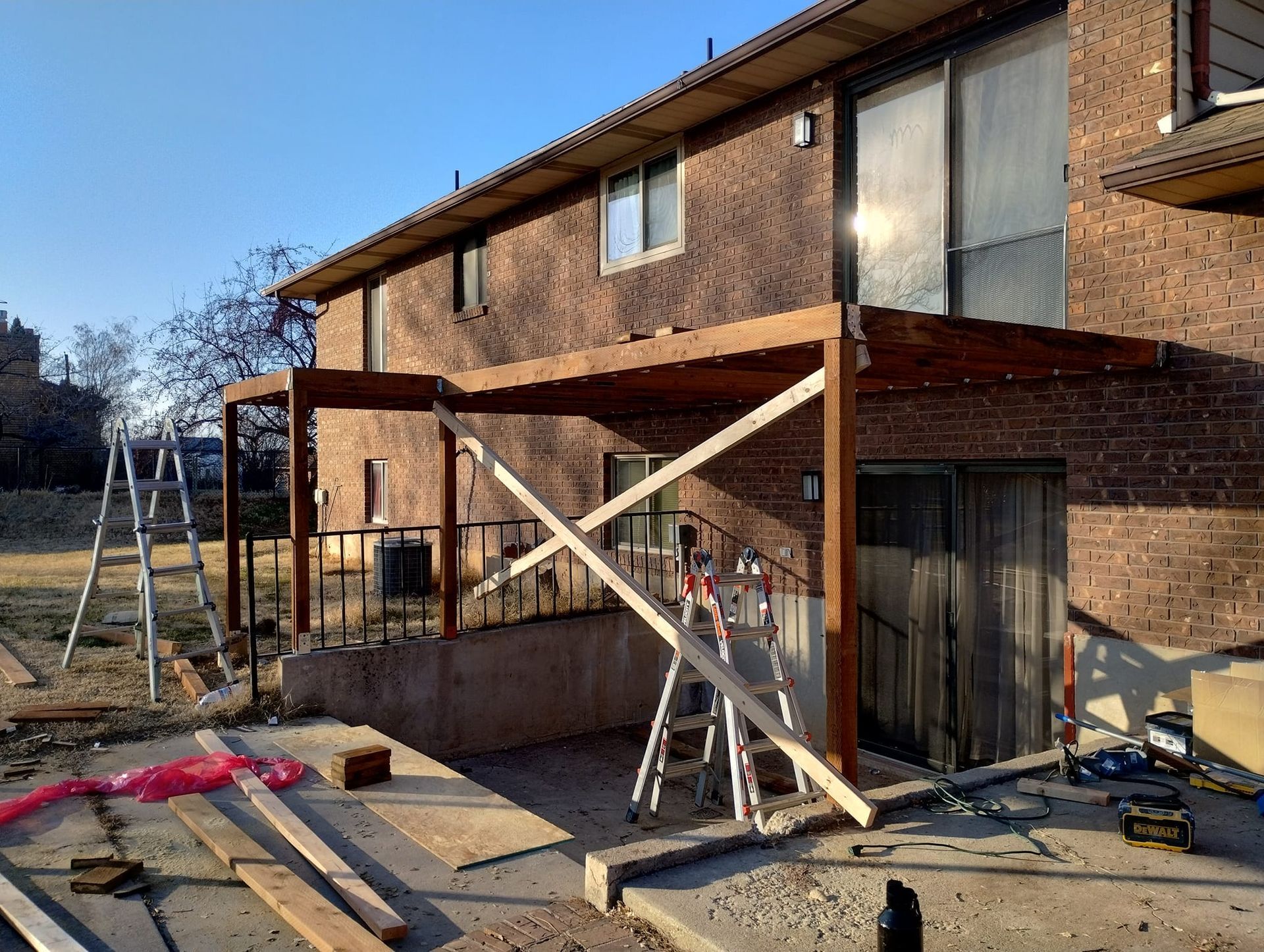 A partially constructed wooden deck frame attached to the back of a brick house with ladders and construction materials.