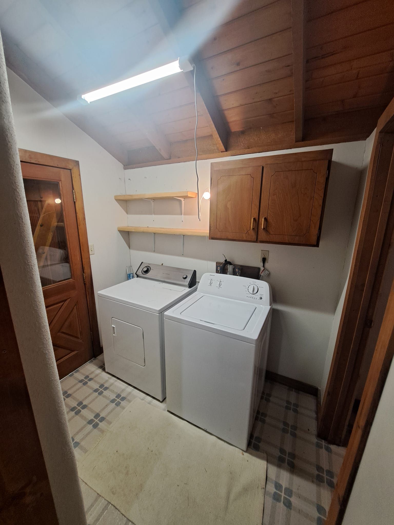 A small laundry room with white washer and dryer, wooden cabinets, two shelves, and a light-colored tiled floor.
