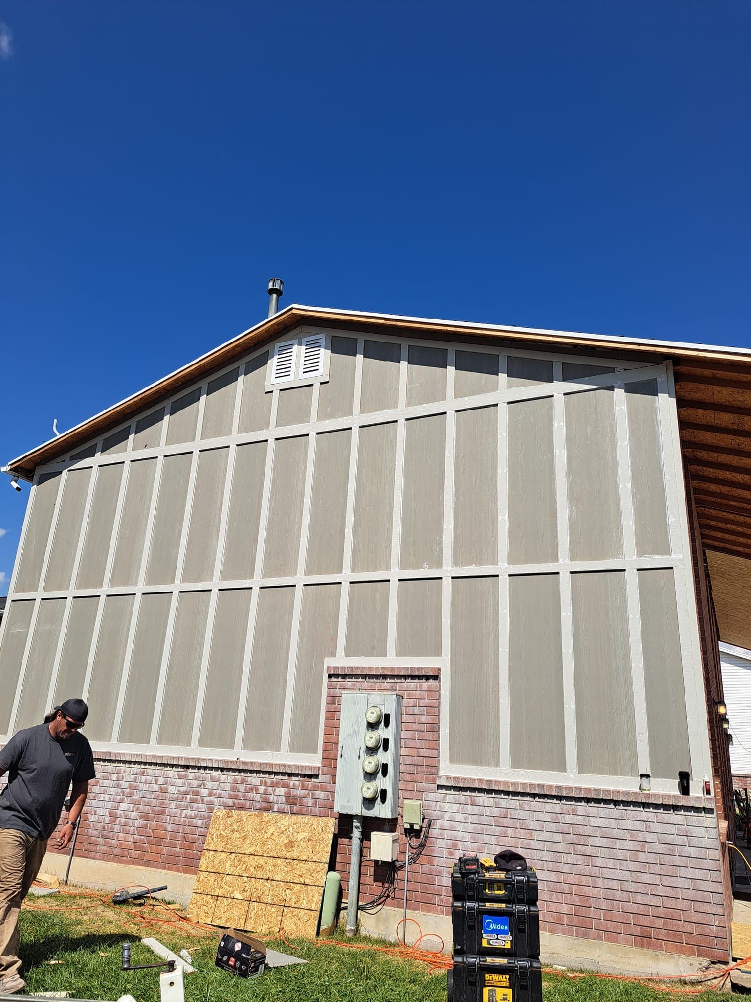A construction worker stands outside a building with gray paneling and an unfinished brick lower wall under a clear sky.