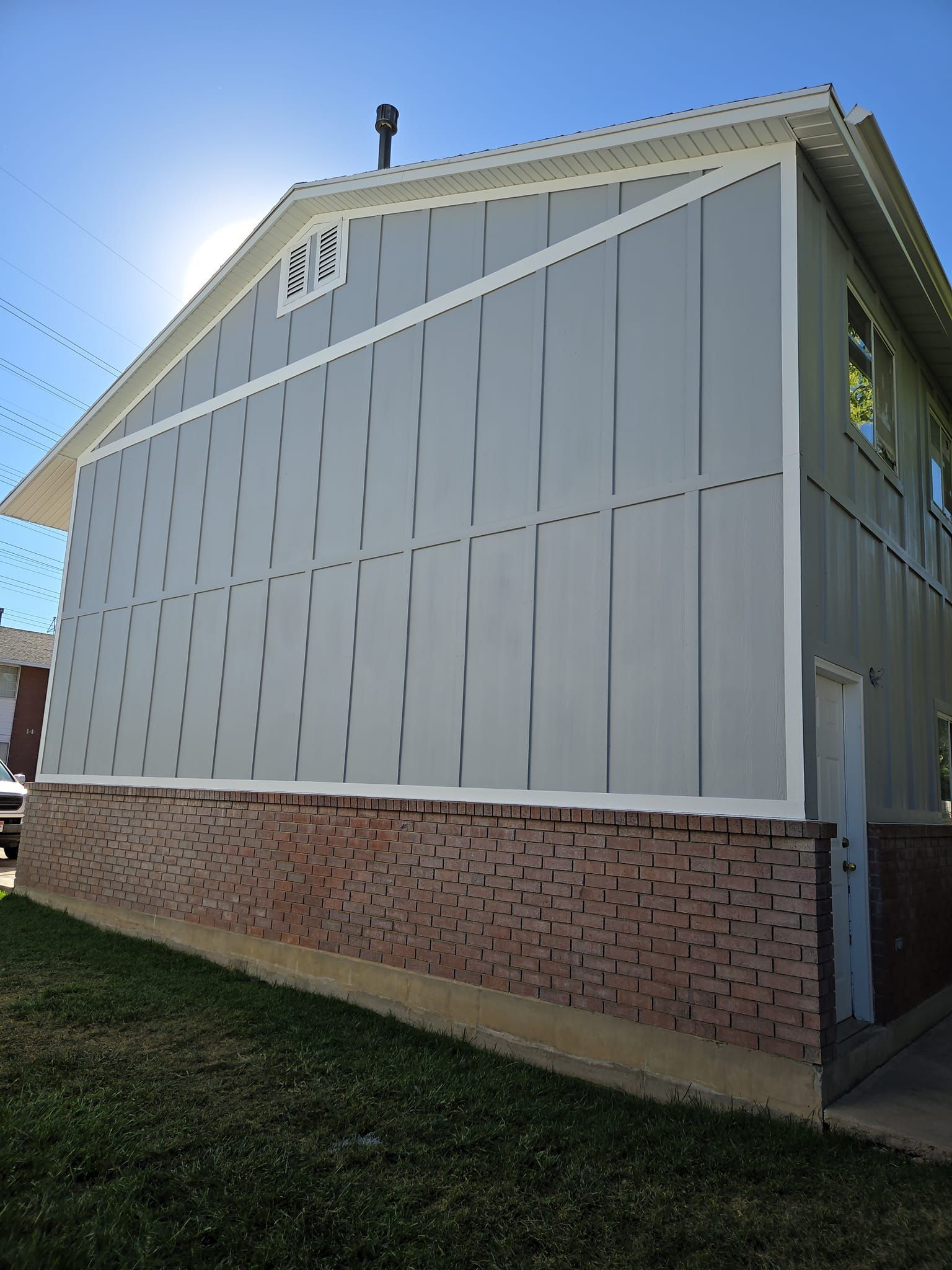Side view of a two-story building with grey vertical board-and-batten siding and a red brick base under a bright blue sky.