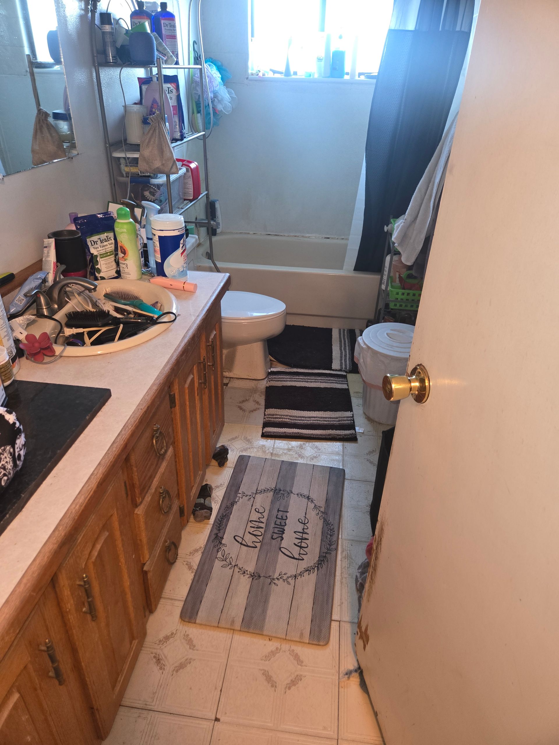 A cluttered bathroom with a wood vanity, sink, toilet, and bathtub, featuring three small rugs on the tiled floor.