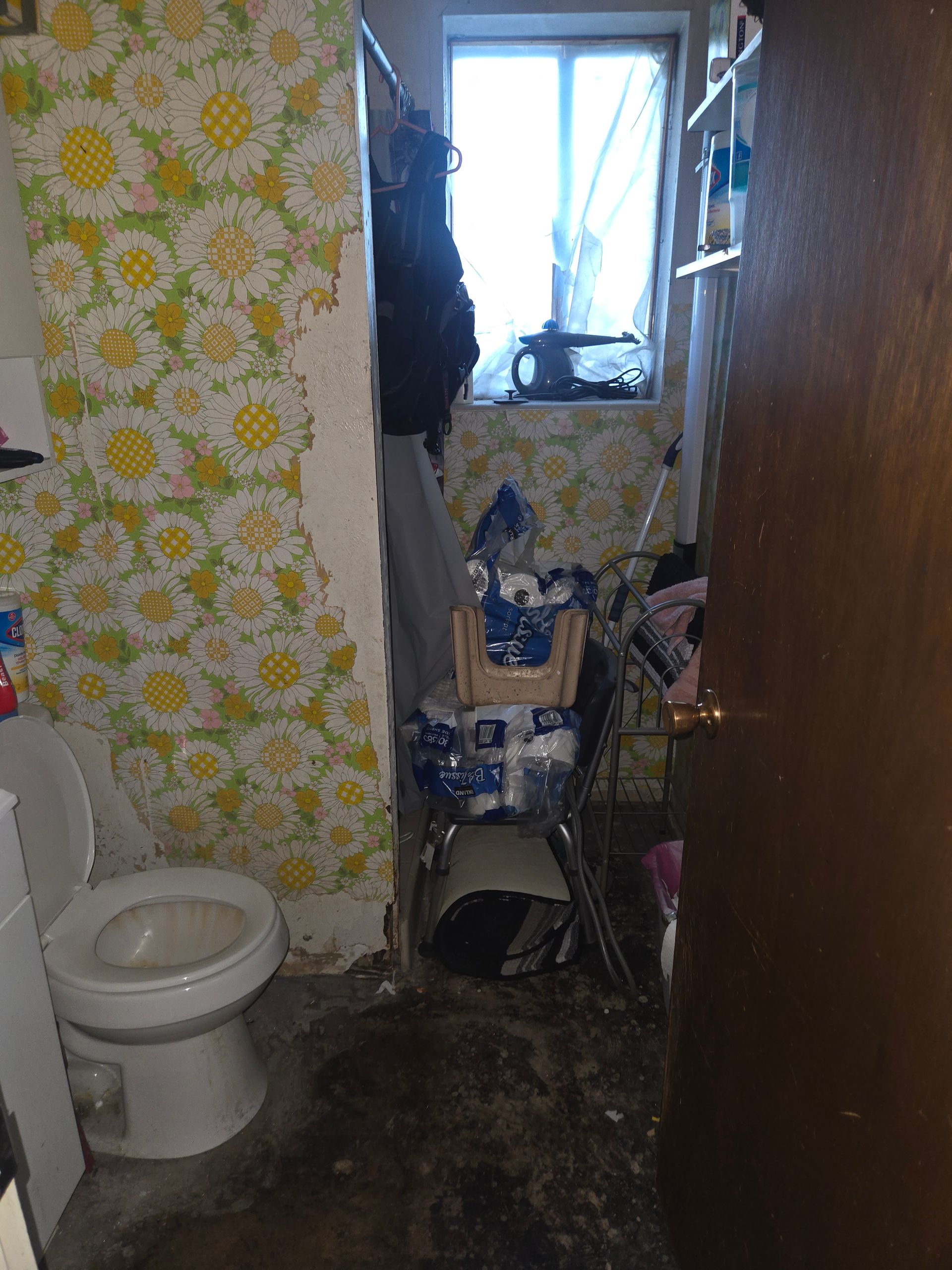 A cluttered bathroom with yellow floral wallpaper, a white toilet, and a dark, stained floor near a window.