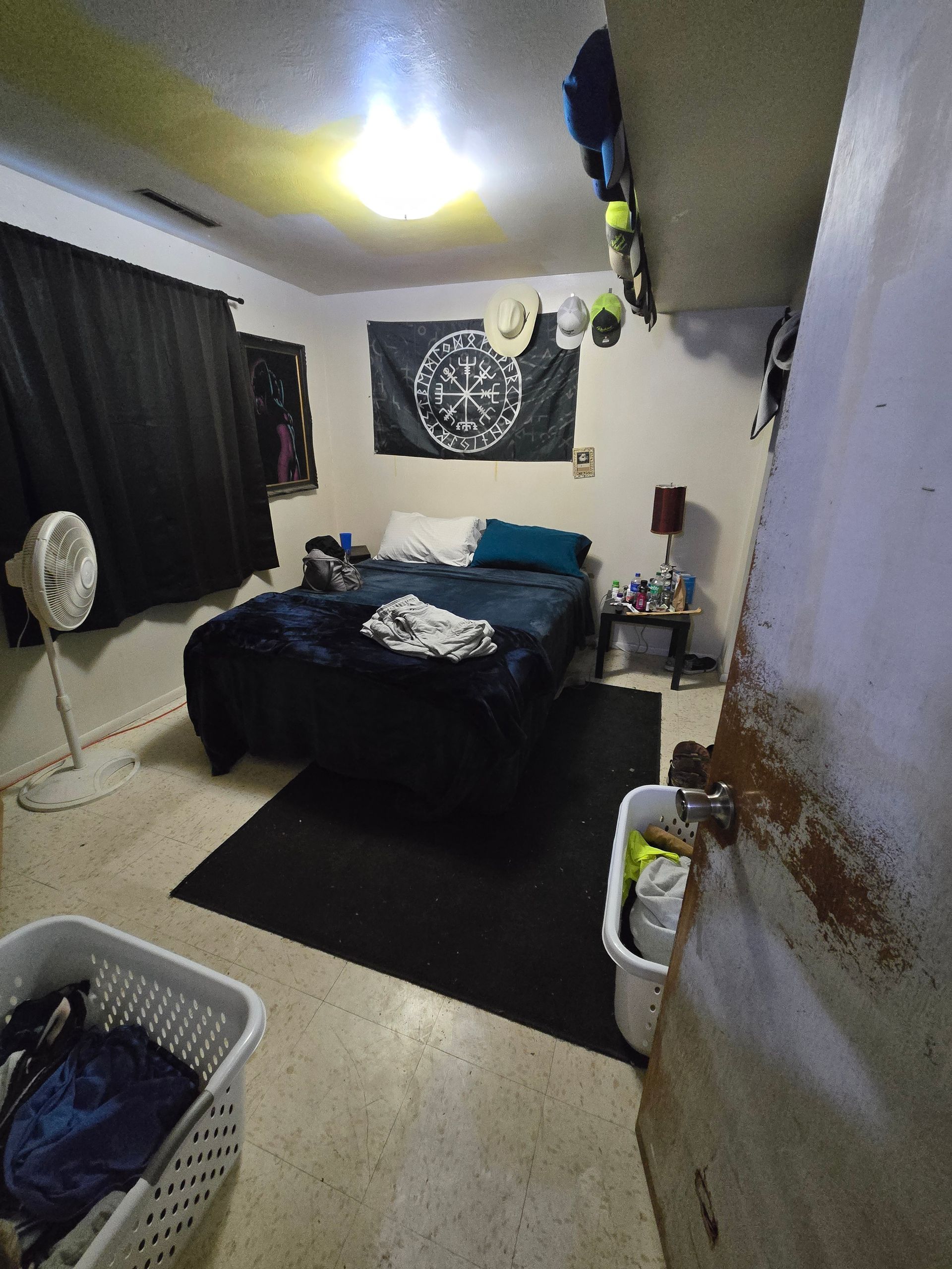 A bedroom with a dark-covered bed, black rug, and laundry baskets on a light-colored floor, viewed through an open doorway.