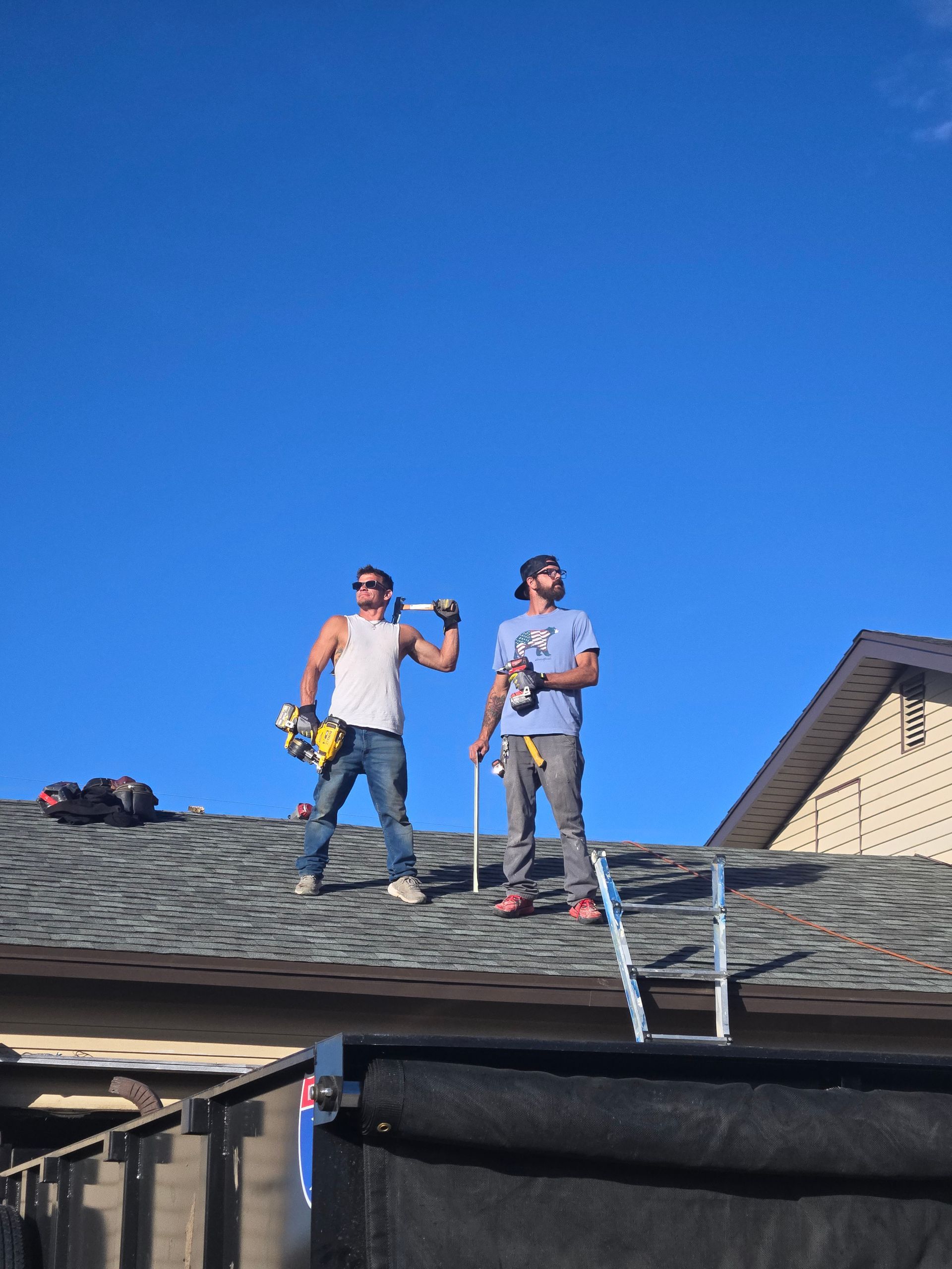 Two workers standing on a roof under a bright blue sky, holding tools and looking to the side.