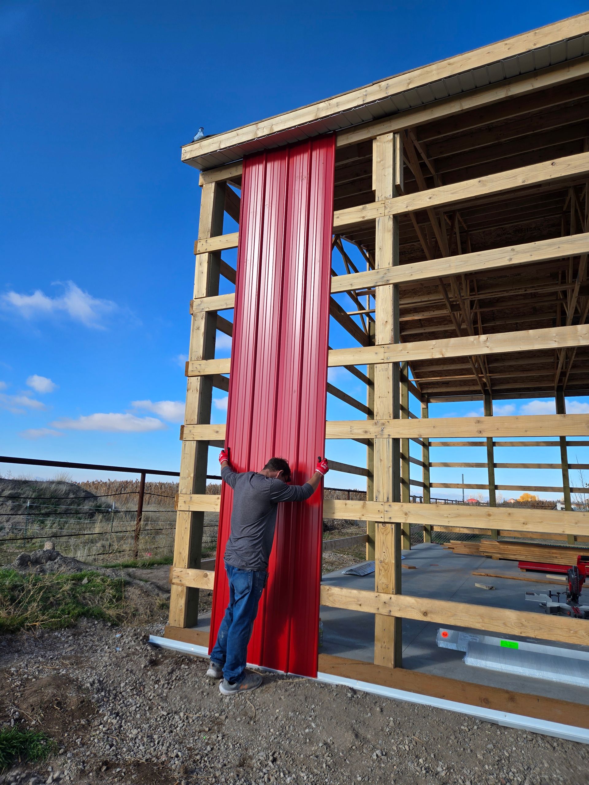 A person installs a vertical sheet of red corrugated metal siding onto the wooden frame of an unfinished barn.