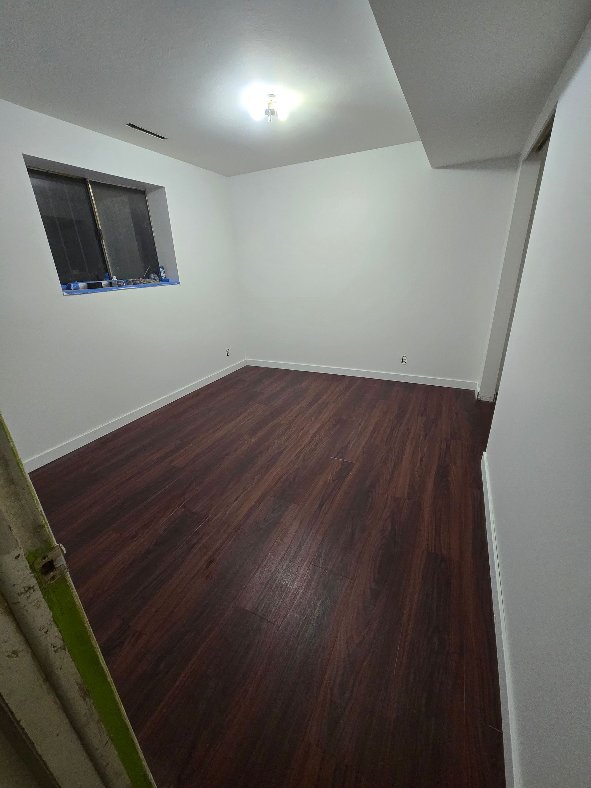 A bright, empty room with white walls, a single window, and dark wood-look laminate flooring.