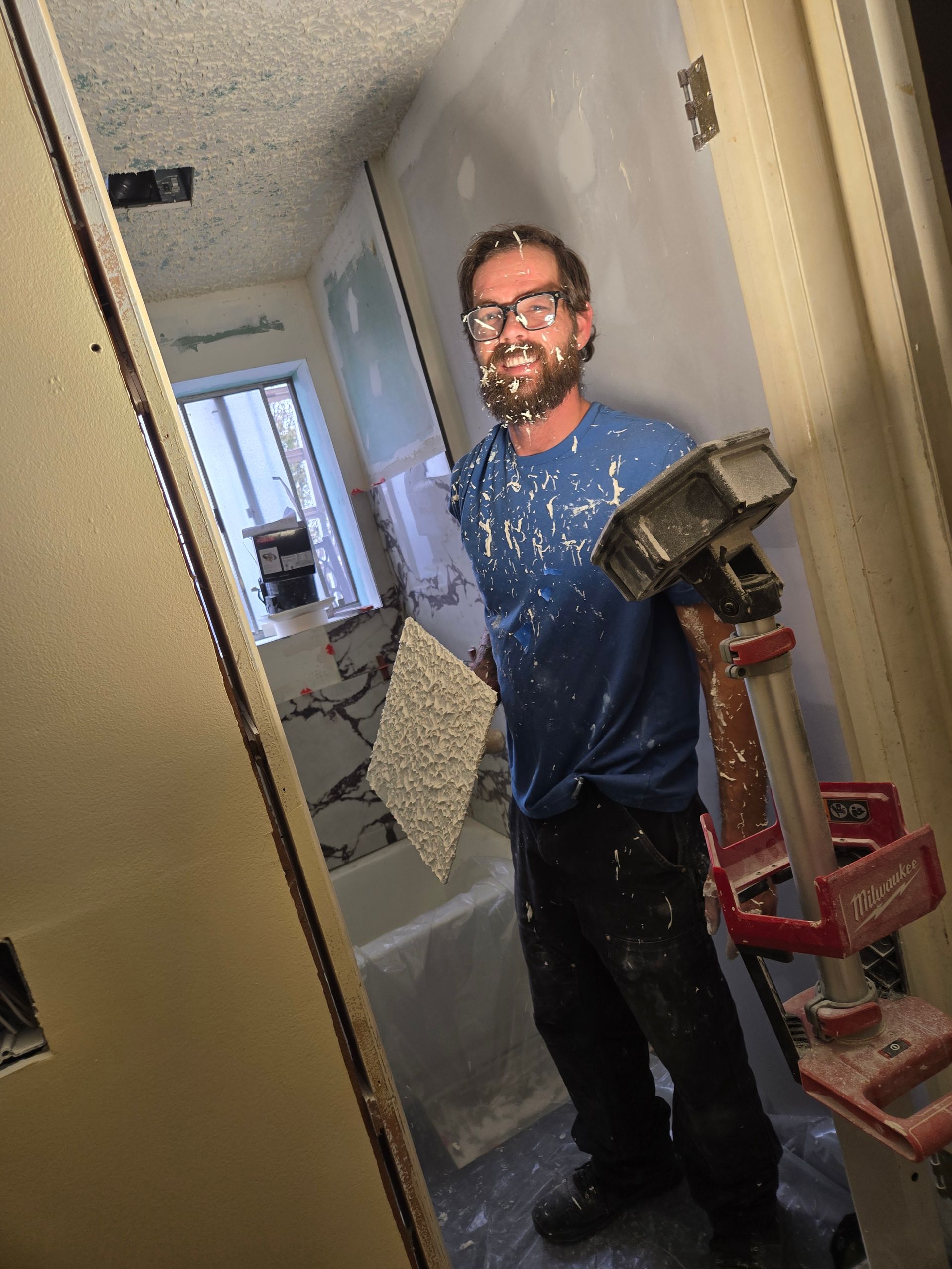 A person covered in drywall mud smiles while working in a bathroom renovation, holding a flat tool in one hand.