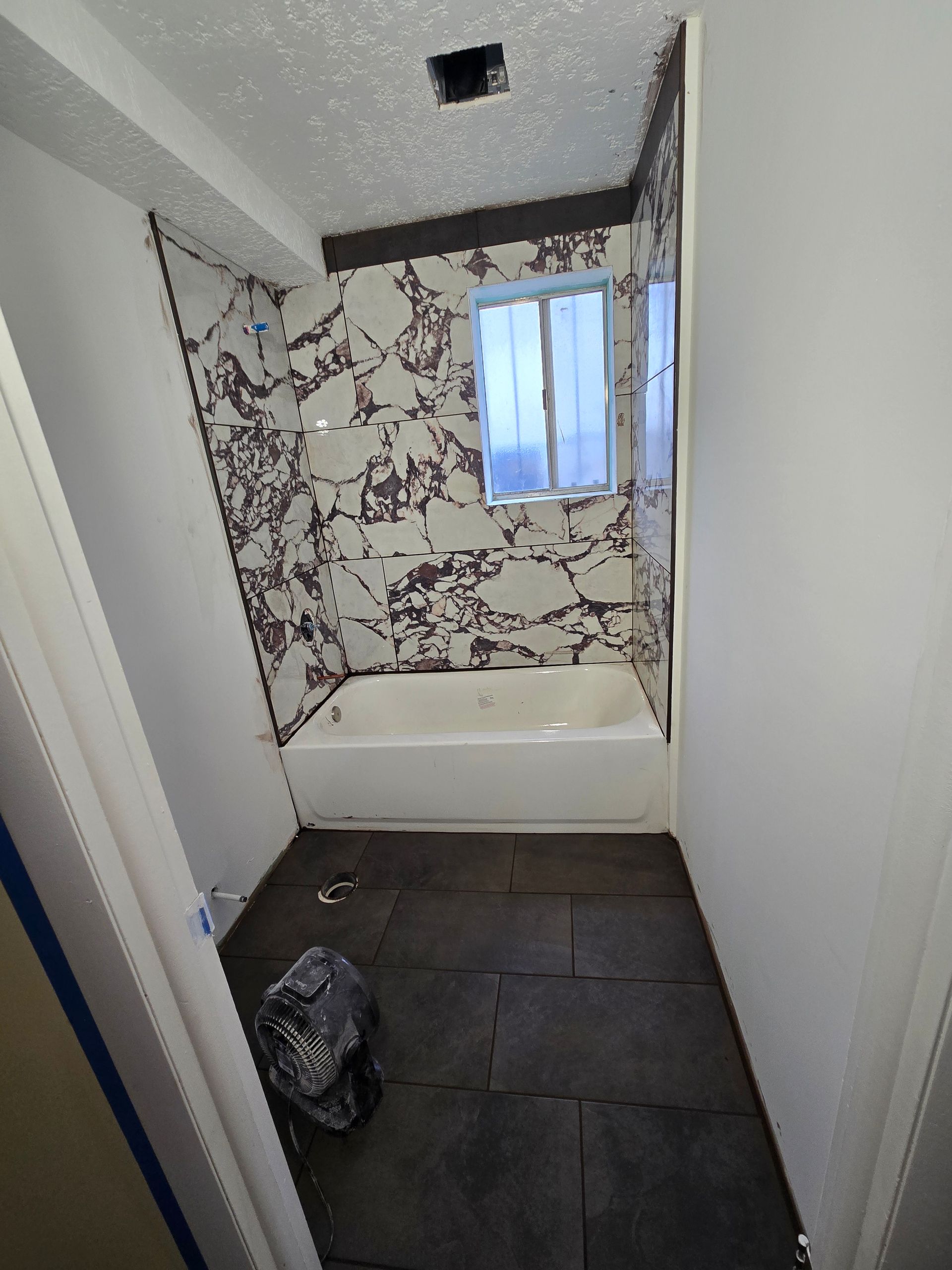 A bathroom with a bathtub, black patterned tile surround, dark floor tiles, and a white ceiling with a small vent.