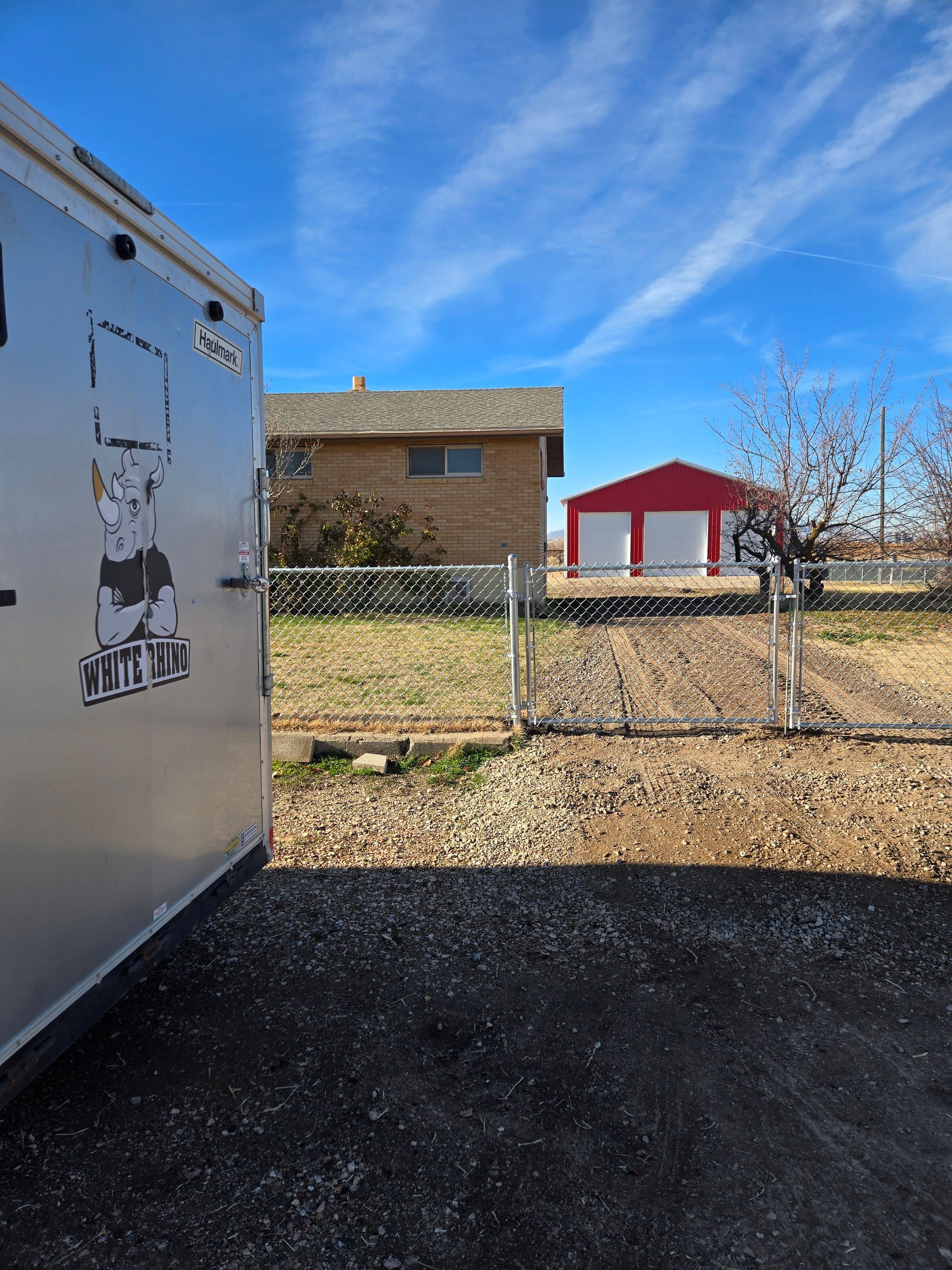 A silver trailer sits in a gravel lot before a fenced-in yard with a house and a red multi-car garage under a blue sky.