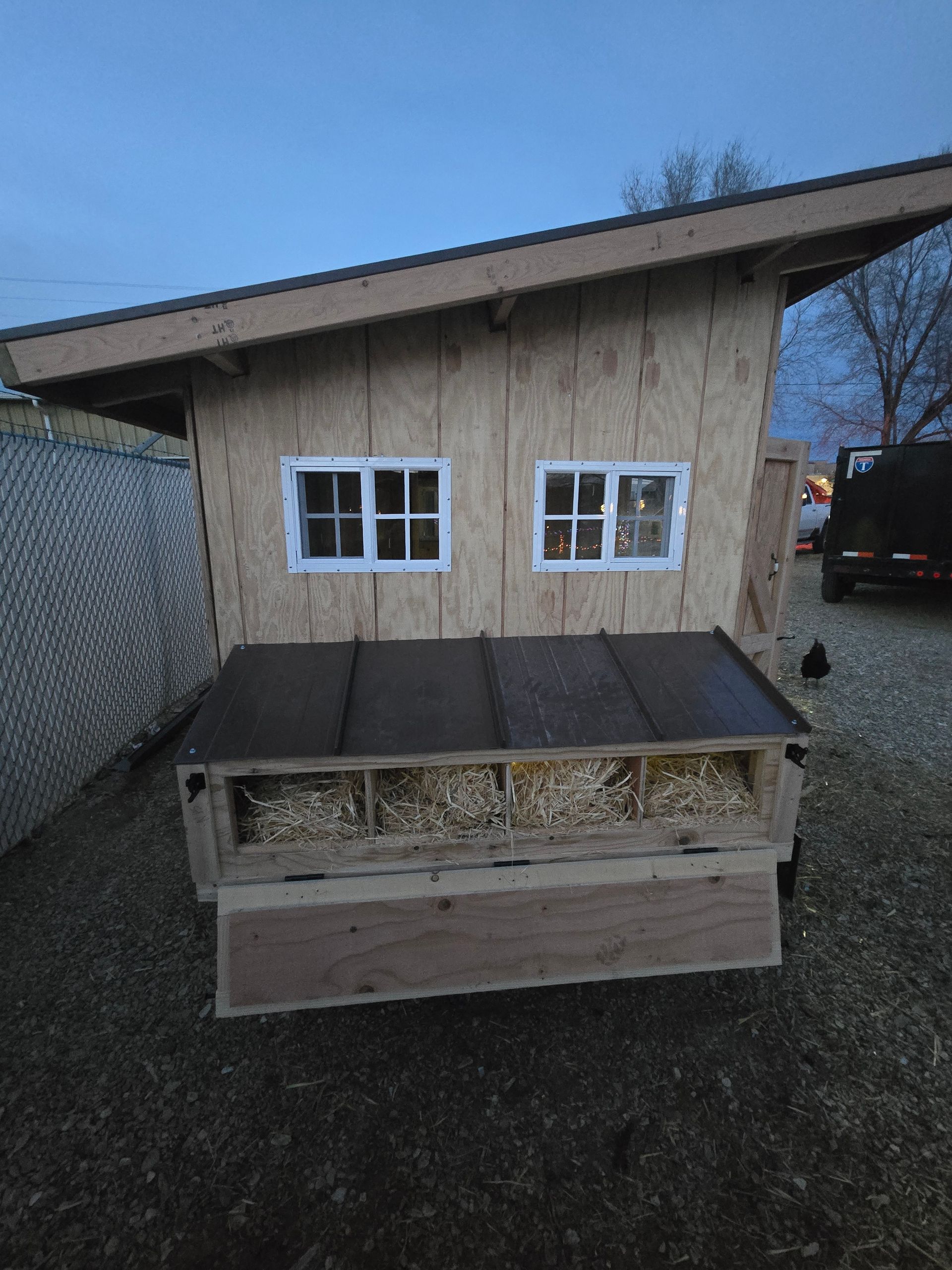 A wooden chicken coop with a slanted roof, two small windows, and an external nesting box filled with wood shavings.