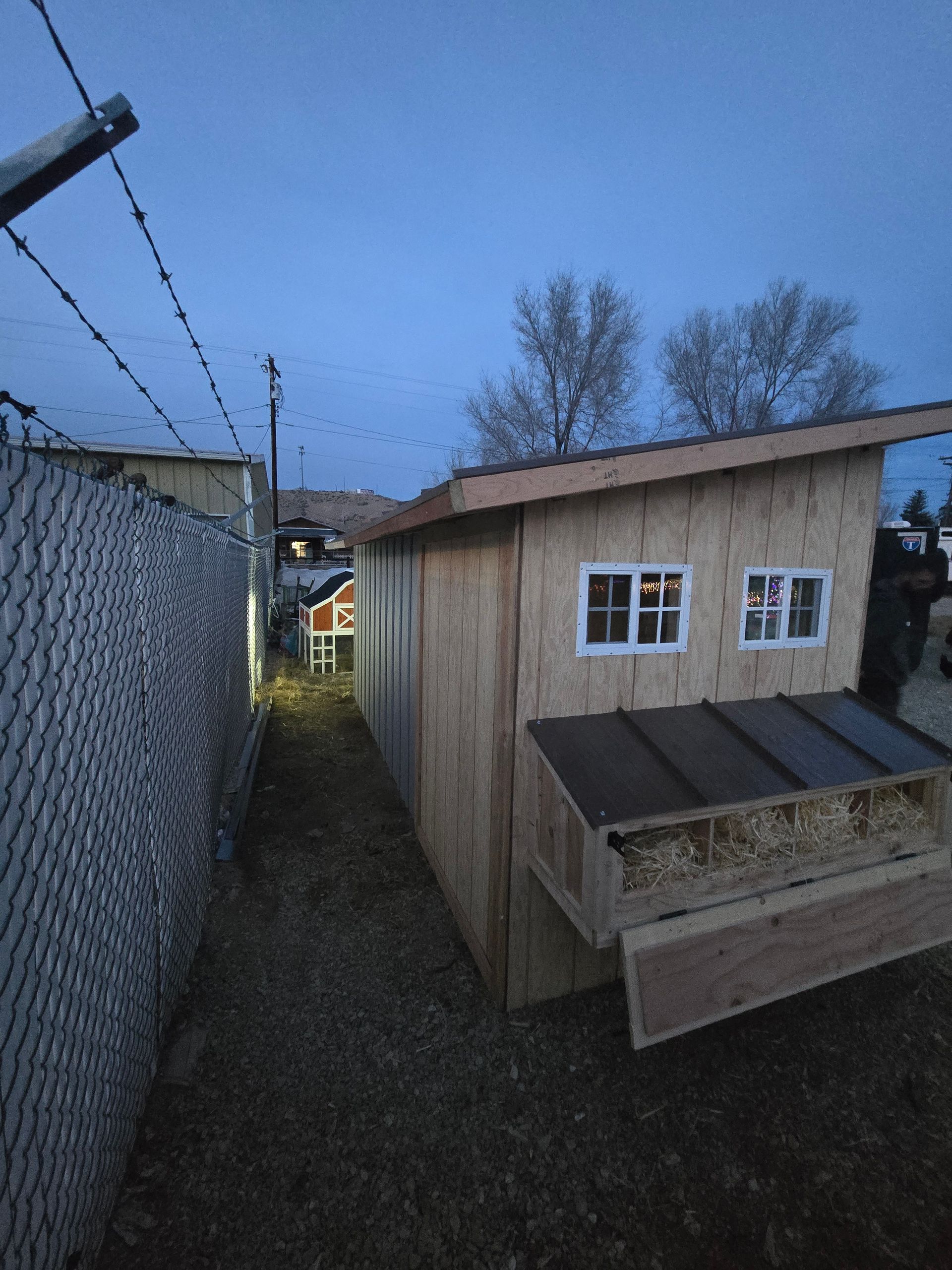 A wooden chicken coop with nesting boxes and small windows sits next to a chain-link fence at dusk.