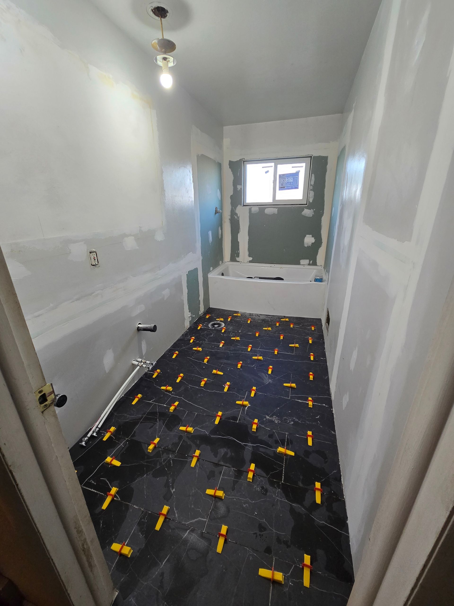 A bathroom under renovation with dark tiled flooring, yellow tile leveling clips, and unpainted drywall walls.