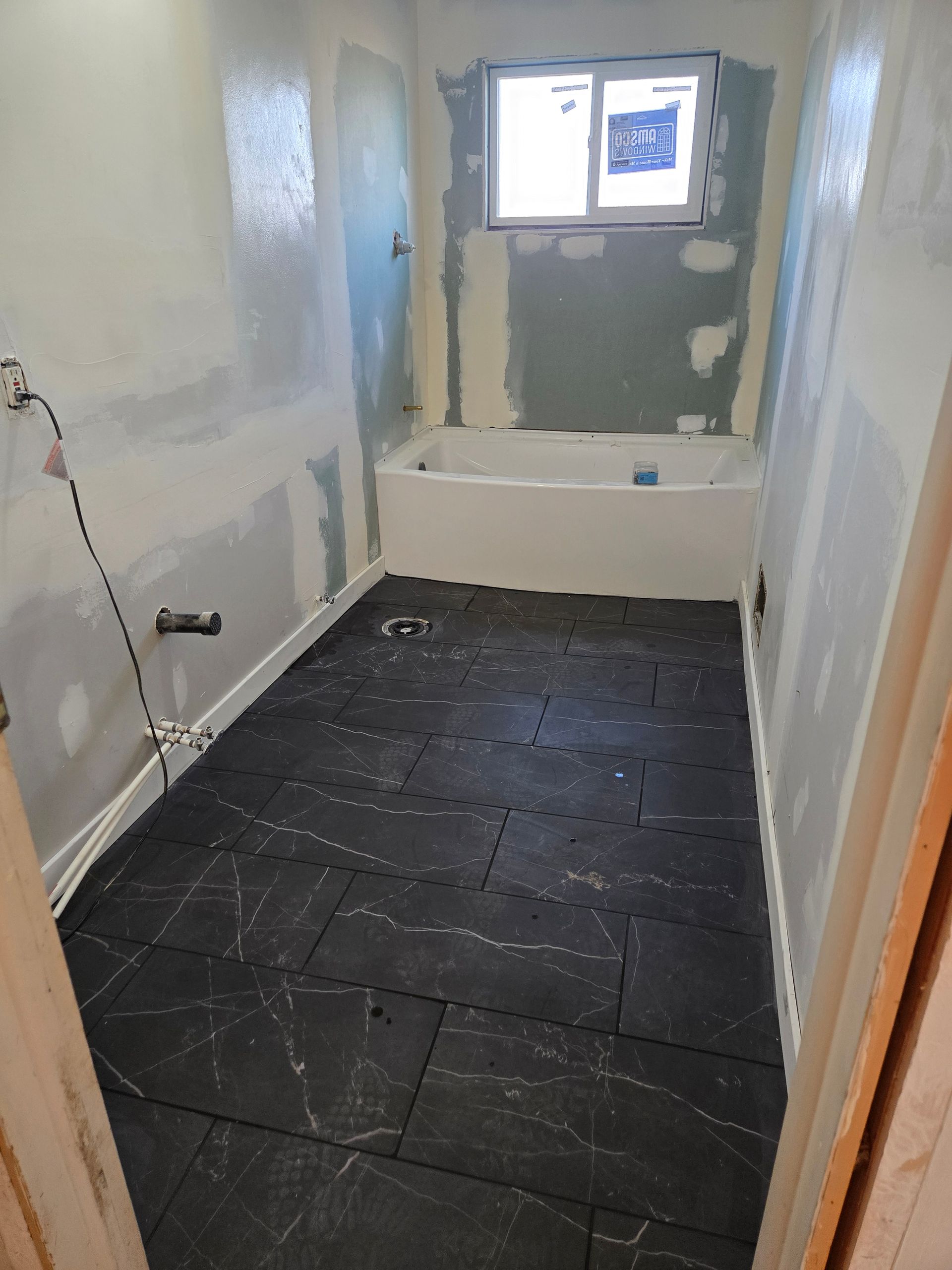 A bathroom under renovation featuring a white bathtub, gray drywall, and black-tiled flooring.