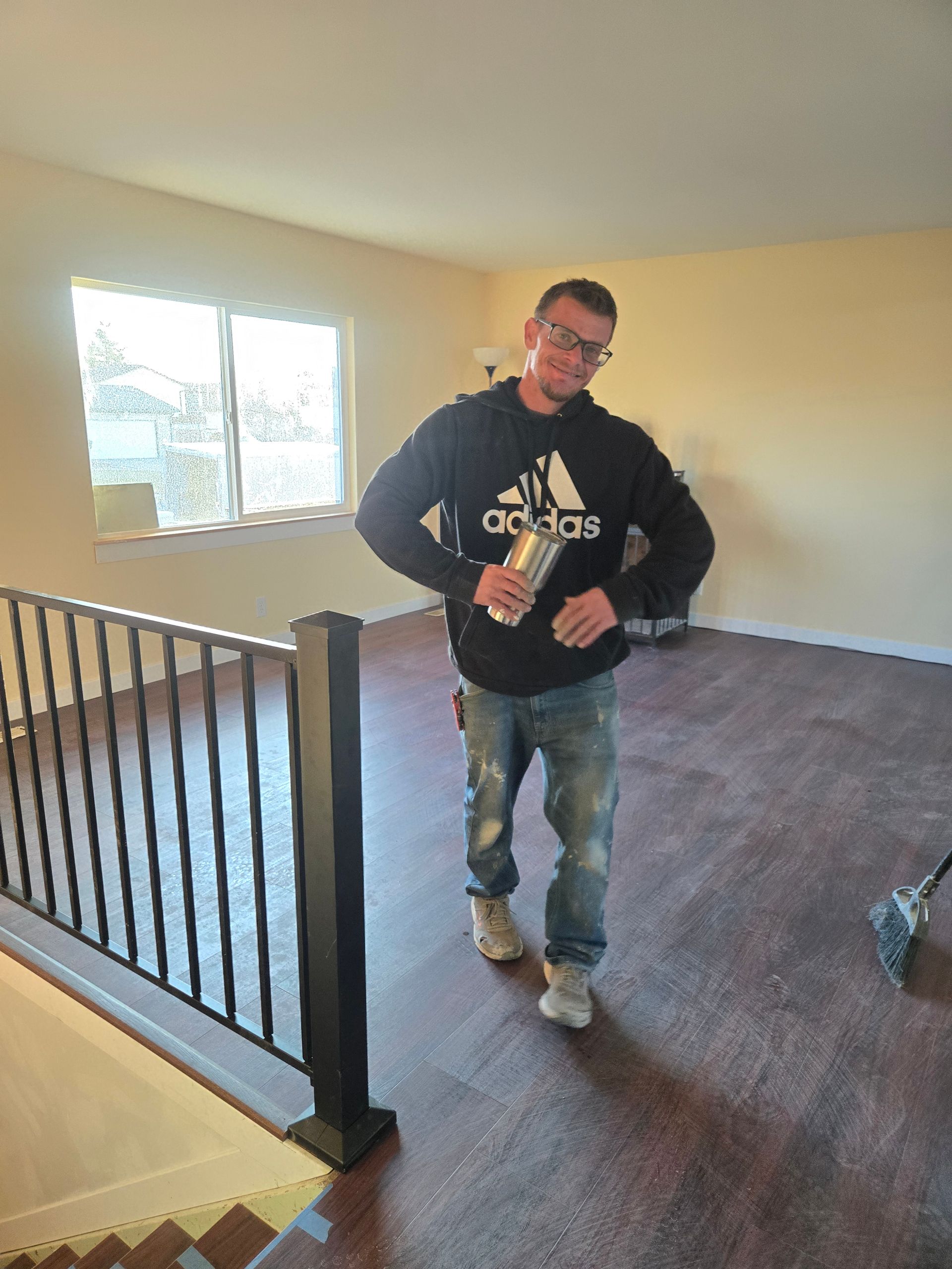 A person in a black Adidas hoodie and jeans stands in an empty, sunlit room with dark wood flooring and a stair railing.
