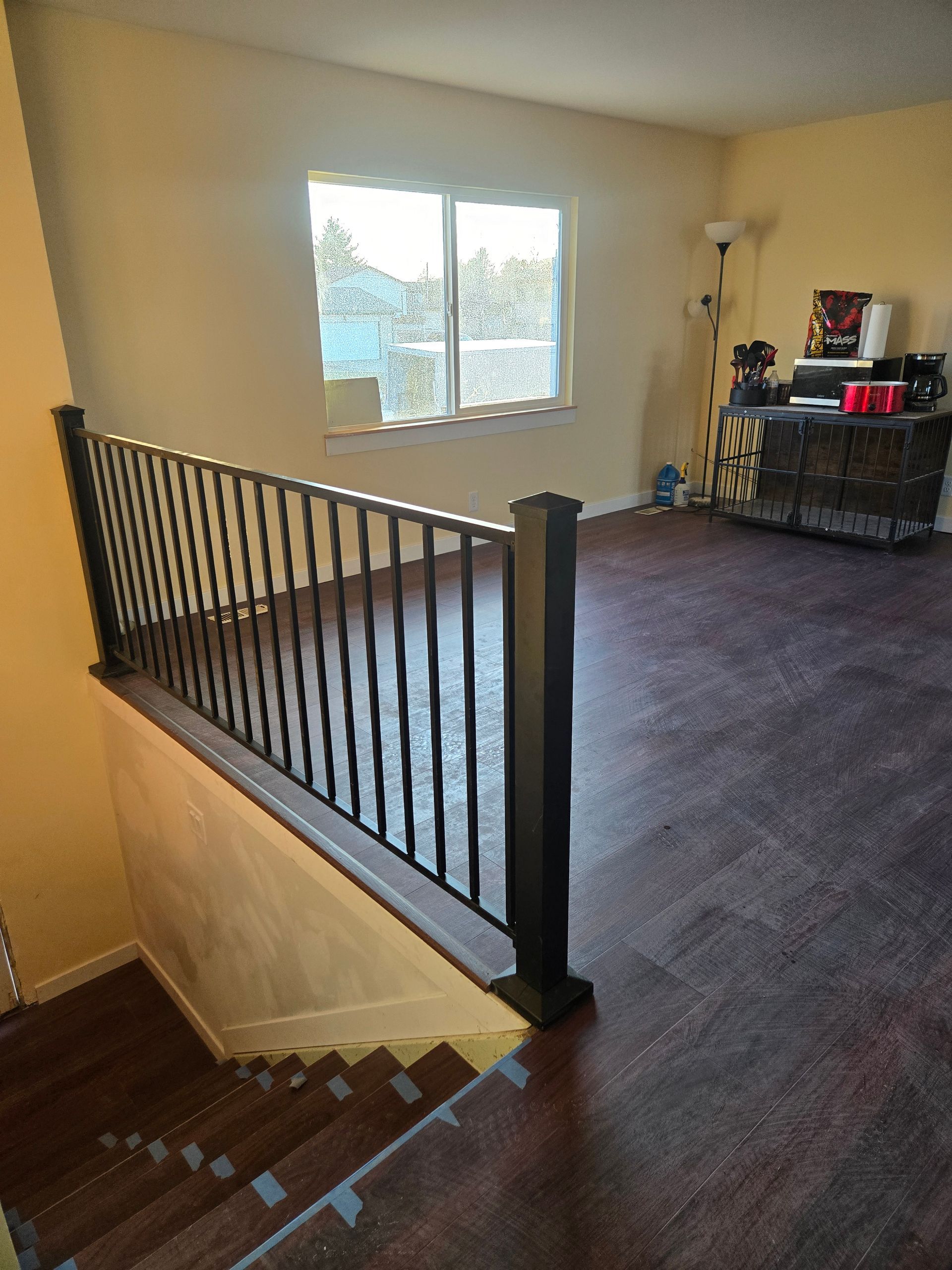 A view from the top of wooden stairs looking into a room with dark wood flooring, a black metal railing, and a window.