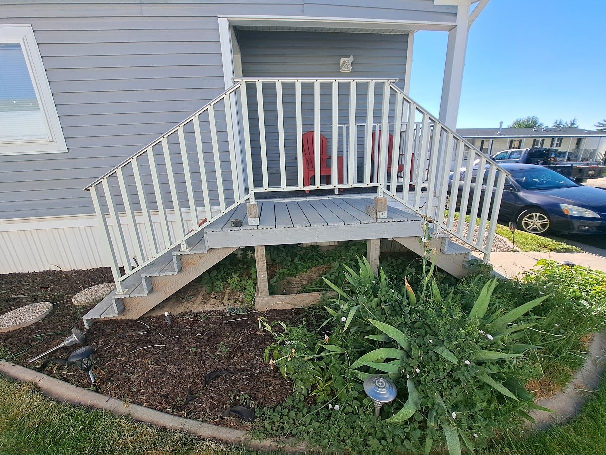 A wooden porch with white railings and stairs leading to the entrance of a gray-sided home, set above a landscaped bed.