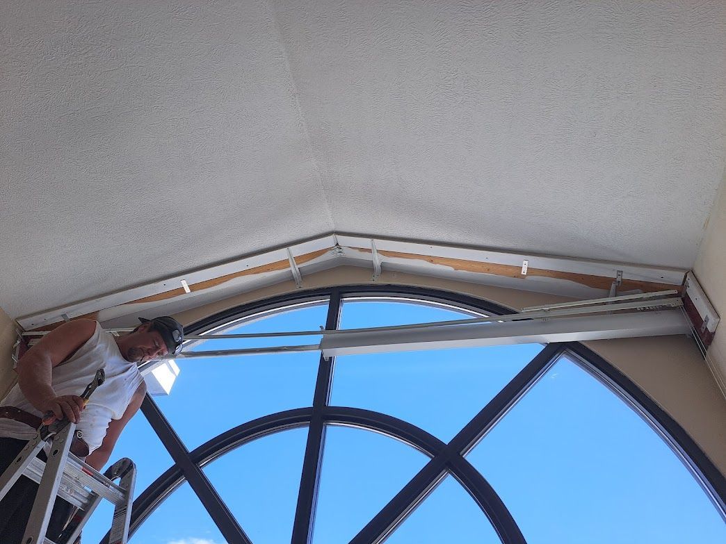 A worker on a ladder installs molding along the top edge of a large arched window.