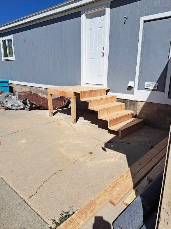 A partially constructed wooden staircase leading to the doorway of a gray residential building on a concrete patio.