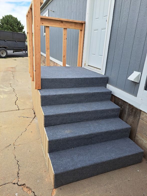A set of five outdoor steps covered in gray carpet with a wooden railing leading to a white door on a gray building.