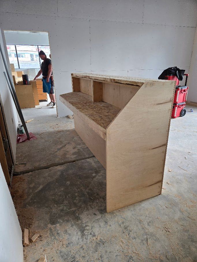 A partially constructed wooden reception desk stands in a room under renovation, with a person working in the background.