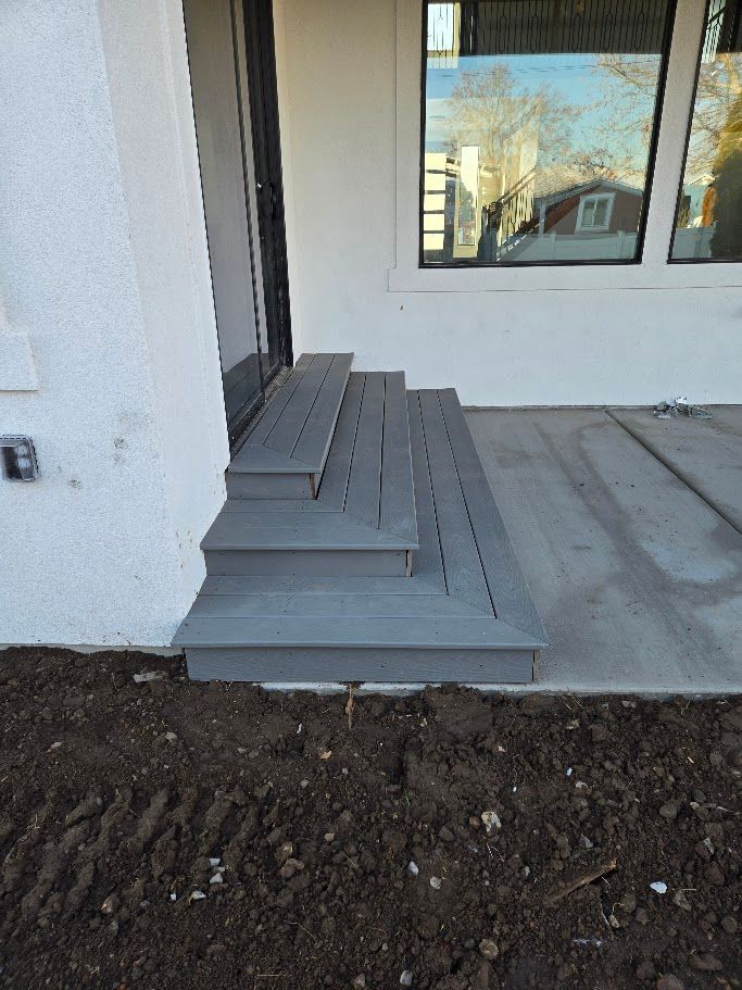 A set of three grey wooden stairs leading up to a glass door on the side of a white house with a dirt patch in the front.