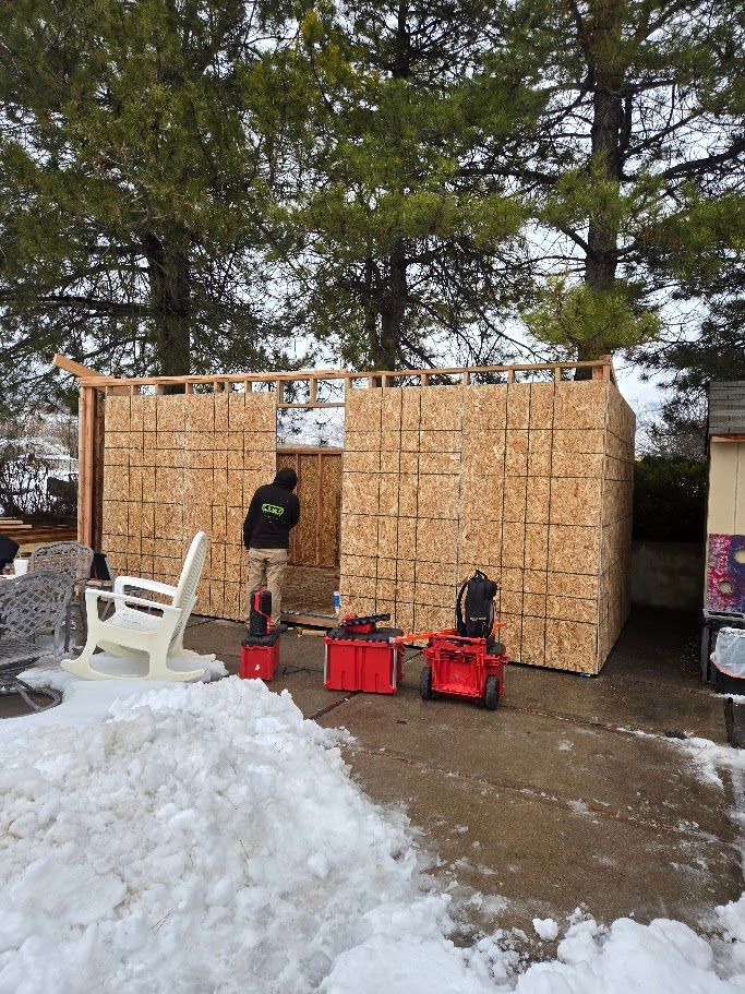 A person in a black jacket installs wooden siding on a shed frame outdoors in the snow with toolboxes nearby.