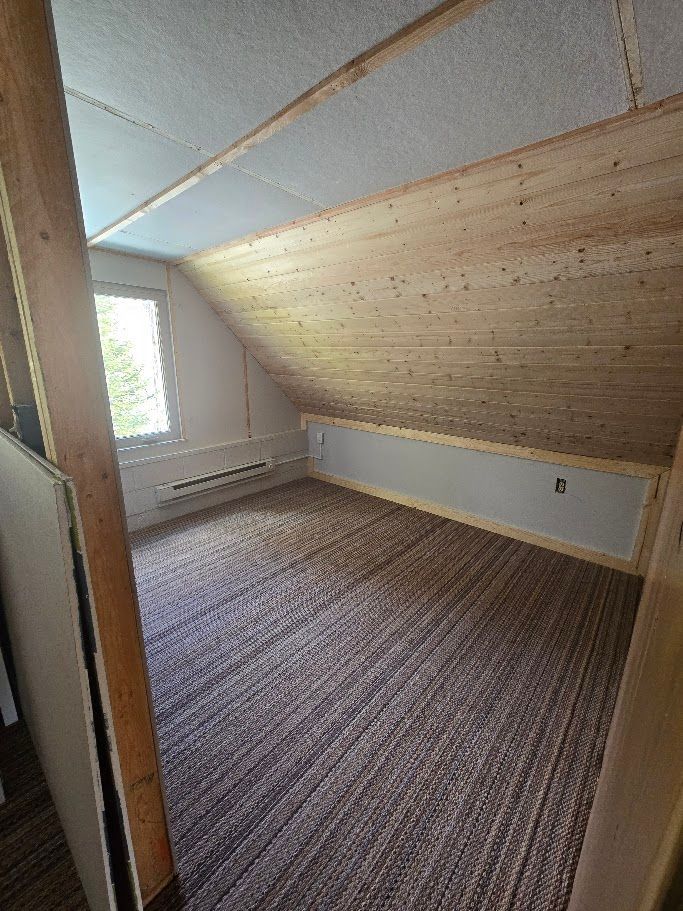 An attic room with a sloped wooden ceiling, brown carpet, light-colored walls, and a window on the left.