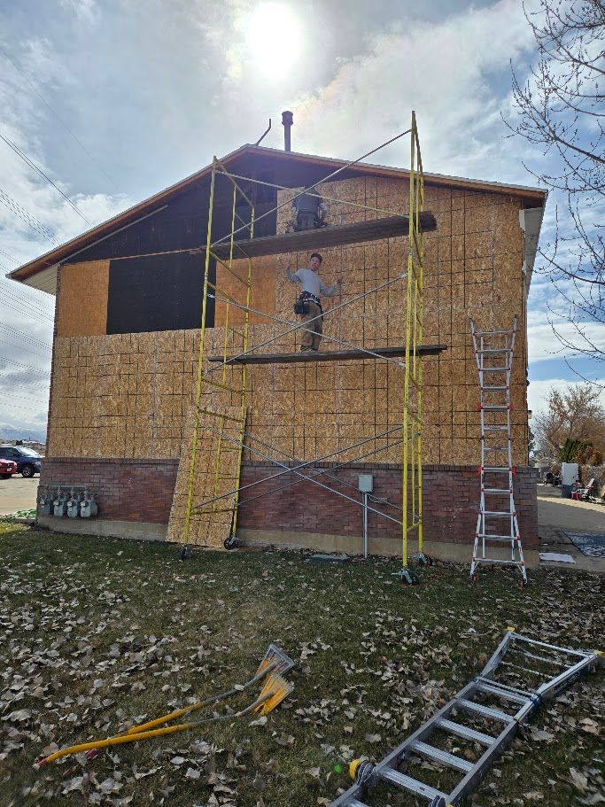 Workers install plywood sheathing on the exterior of a two-story house using yellow metal scaffolding.