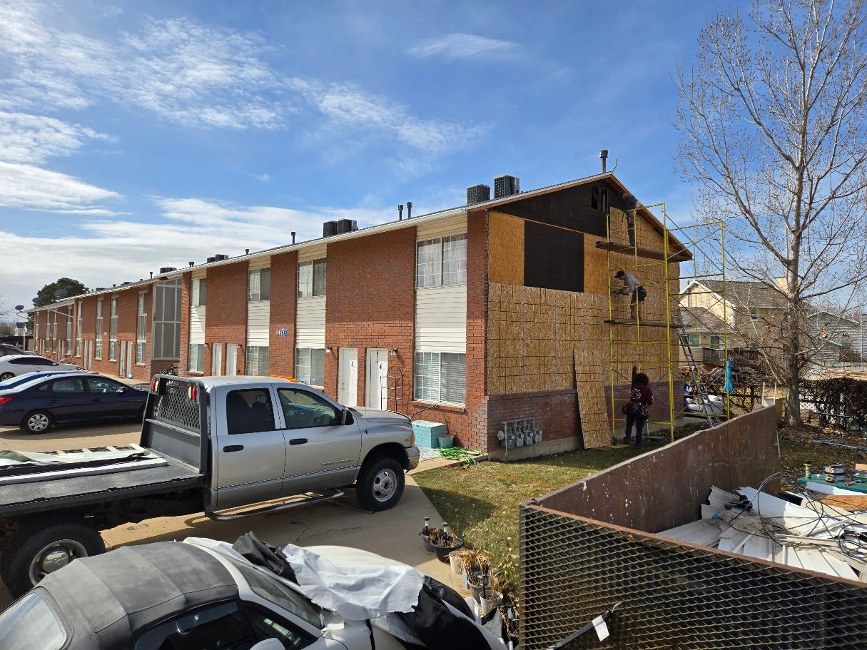 Workers repair the plywood exterior of a two-story brick apartment building on a sunny day.