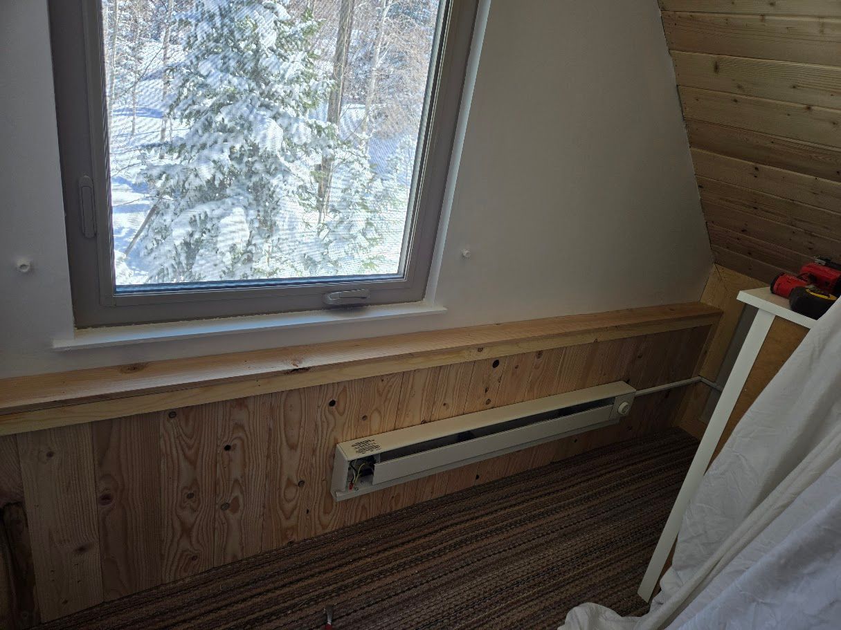 A window looks out onto a snowy forest above a wooden wall with an electric baseboard heater.